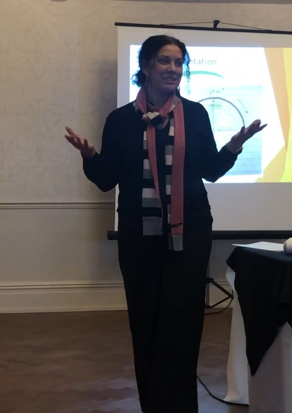 A woman standing in front of a presentation screen, gesturing with both hands, wearing a black blazer, black pants, and a striped scarf, during a presentation.