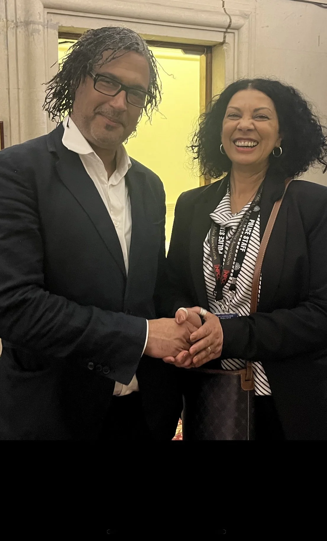 Two people shaking hands and smiling at the camera, one man in a dark suit with glasses and an open-collared shirt, and one woman with dark curly hair wearing a suit and police staff badge.