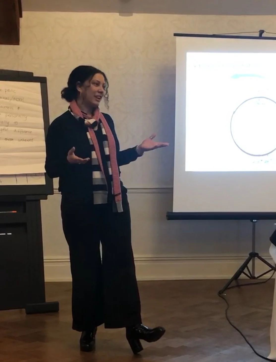 A woman giving a presentation indoors, standing near a projector screen displaying a slide with a circle. She is wearing a black outfit, a striped scarf, and black platform shoes. She has dark, curly shoulder-length hair and is gesturing with her hands.
