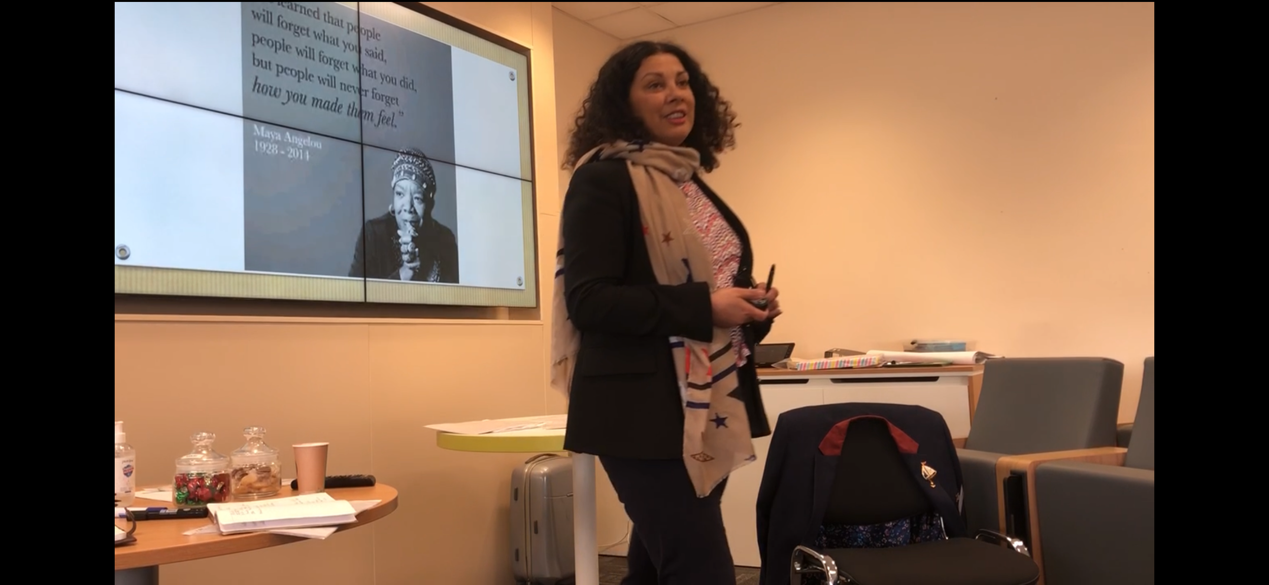 A woman with curly hair wearing a black blazer and a beige scarf with stars is standing in a conference room, holding a remote and giving a presentation. Behind her, a screen displays a slide featuring a quote and a photo of Maya Angelou with the years 1928-2014. The room has chairs, a table with various items including jars, papers, and a cup, and a gray wall.