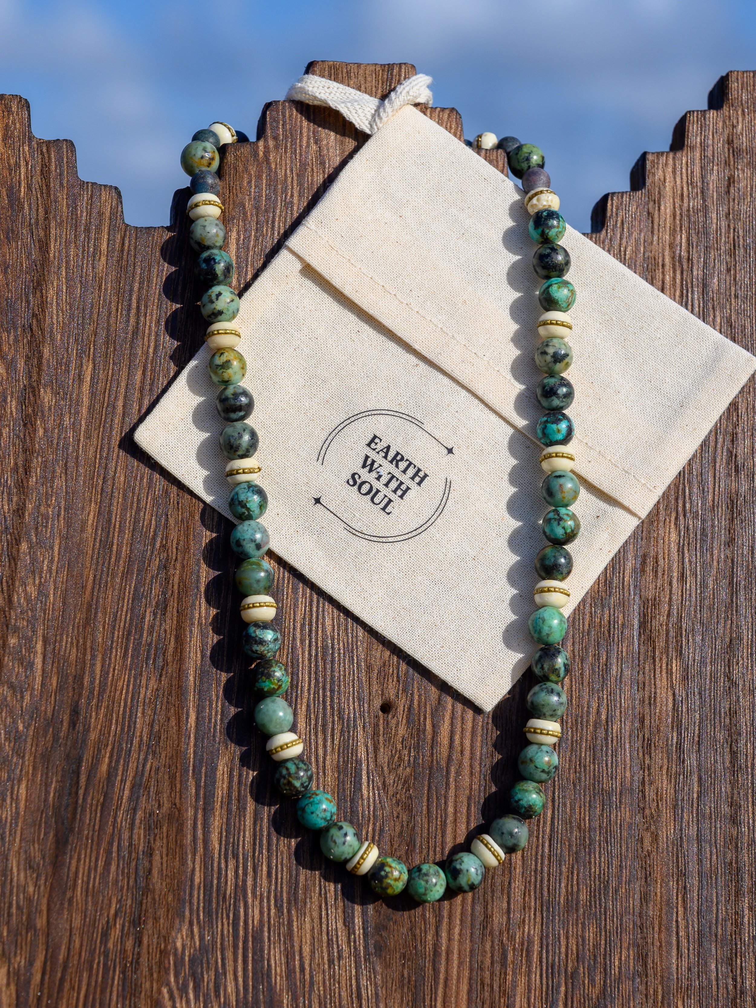 A beaded necklace with green, black, and cream-colored round beads and gold spacer beads, draped over a beige cloth and a wooden surface with a blue sky background and a wooden fence.