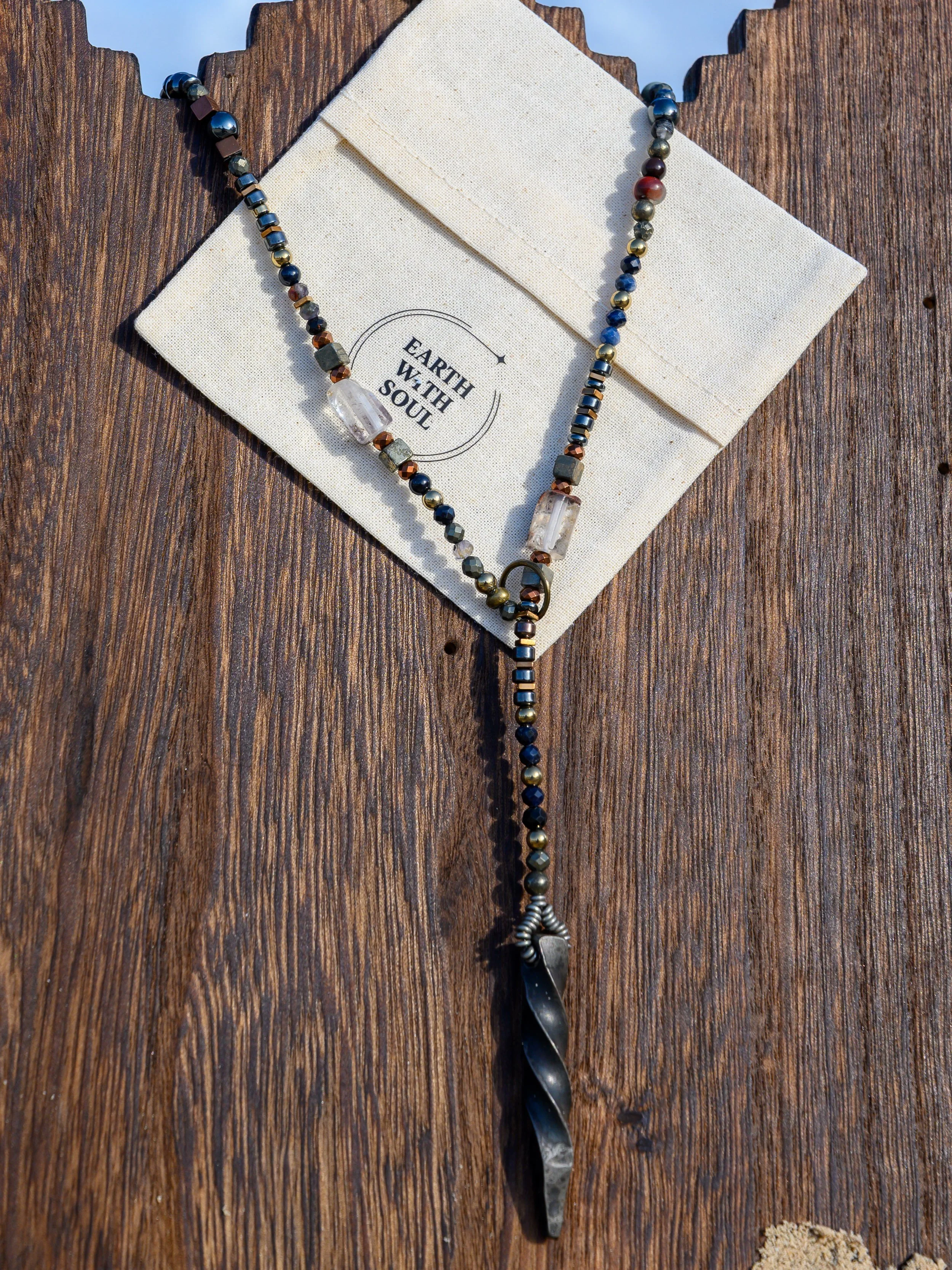 Beaded necklace with metallic and stone beads, featuring a spiked metal pendant, lies on a white cloth with the words "Earth with Soul" printed on it. The cloth is placed on a wooden surface.