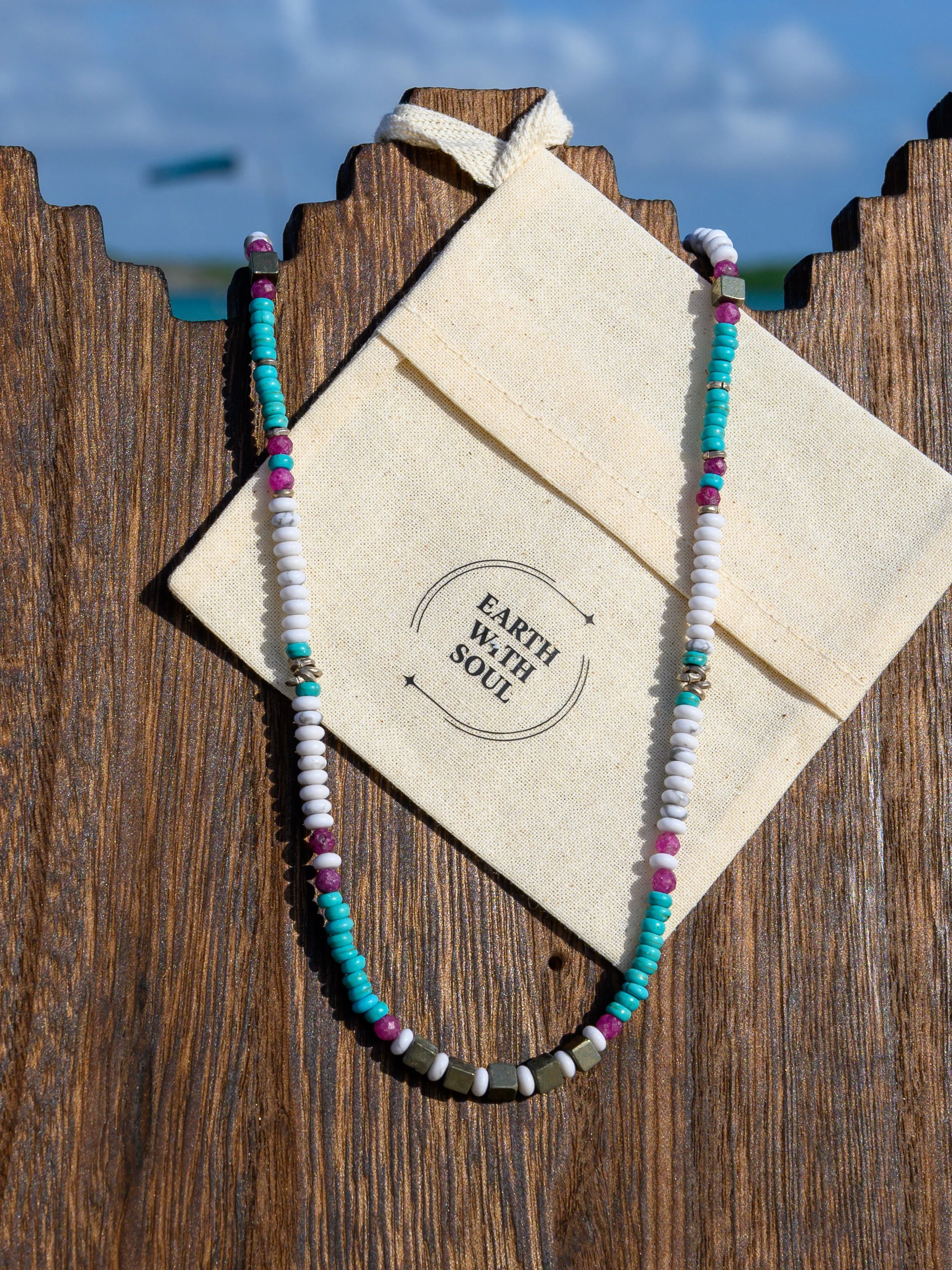 Colorful beaded necklace draped over a beige envelope with the text "Earth W. the Soul" printed on it, on a wooden surface with a cloudy sky in the background.