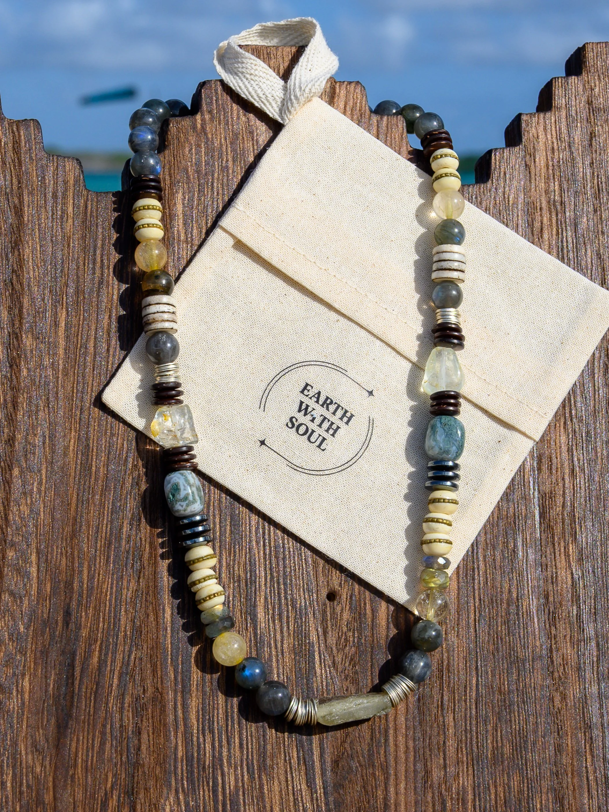 Beaded necklace with various gray, white, brown, and yellow beads, laying on a beige cloth and wood surface, with a pocket-sized card that has the words "Earth With Soul" printed on it.