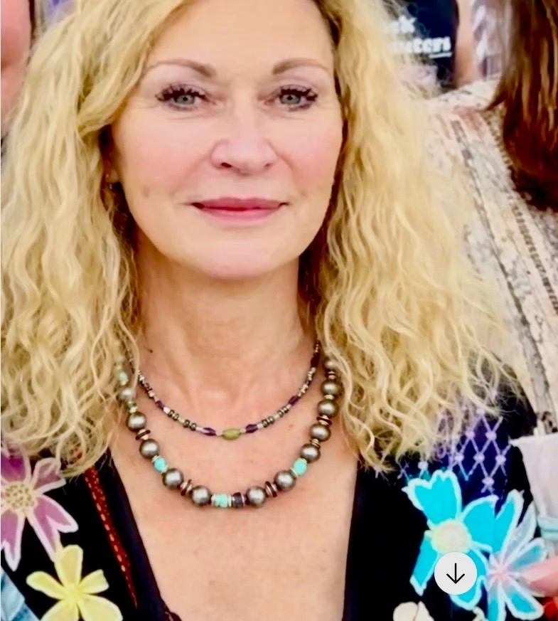 Close-up of a woman with long curly blonde hair, wearing layered necklaces and a floral-patterned top.