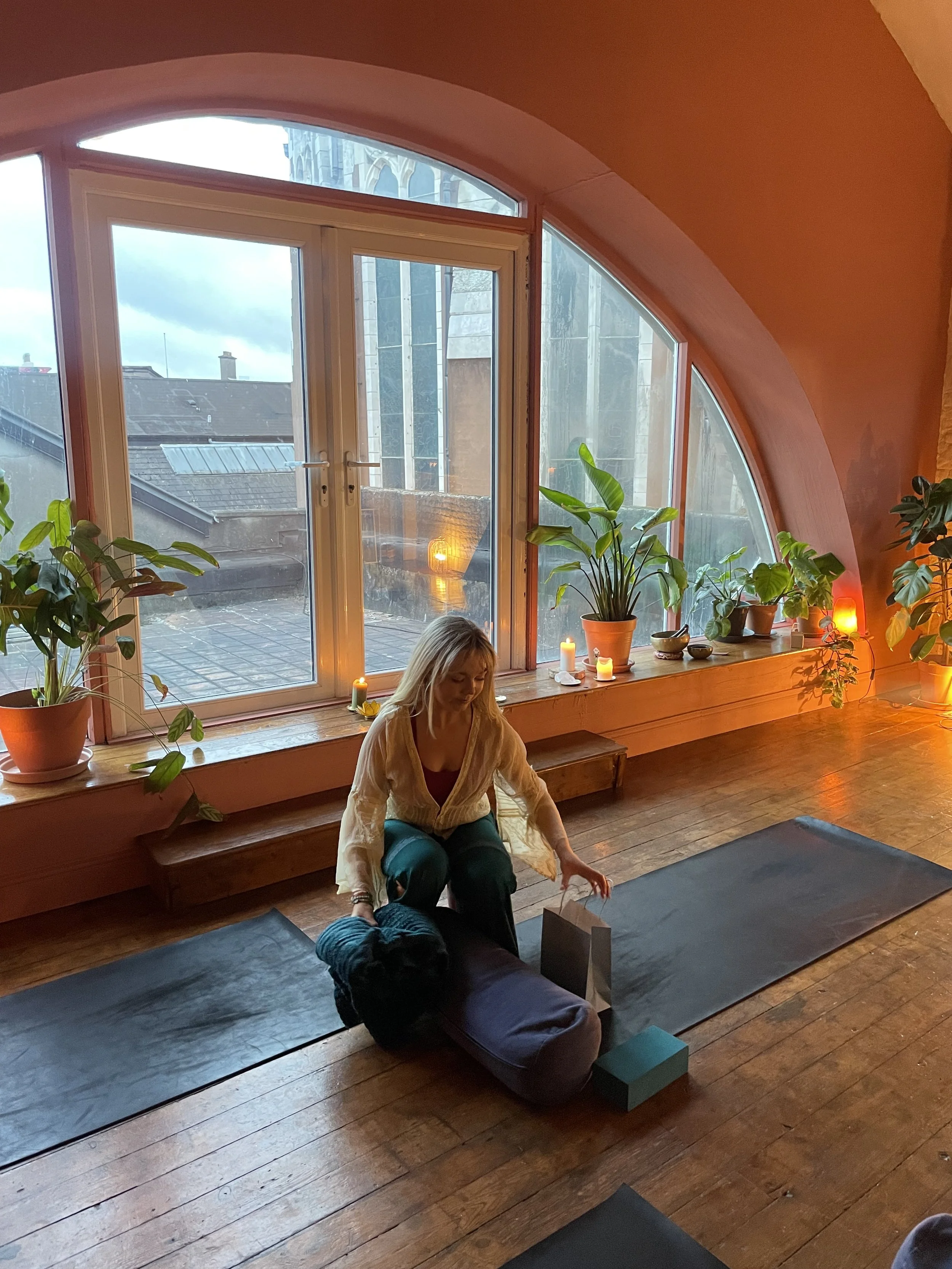 A woman practicing yoga indoors on a black yoga mat, with a large arched window, potted plants, candles, and wooden flooring.
