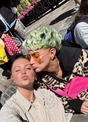 Mother and Daughter taking a selfie outdoors on a city street, one with light green curly hair and wearing orange-tinted sunglasses, and the other with her eyes closed, puckering her lips.