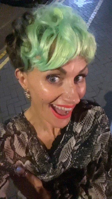 A mother with short, curly hair dyed bright green and blonde, smiling and taking a selfie outdoors at night.