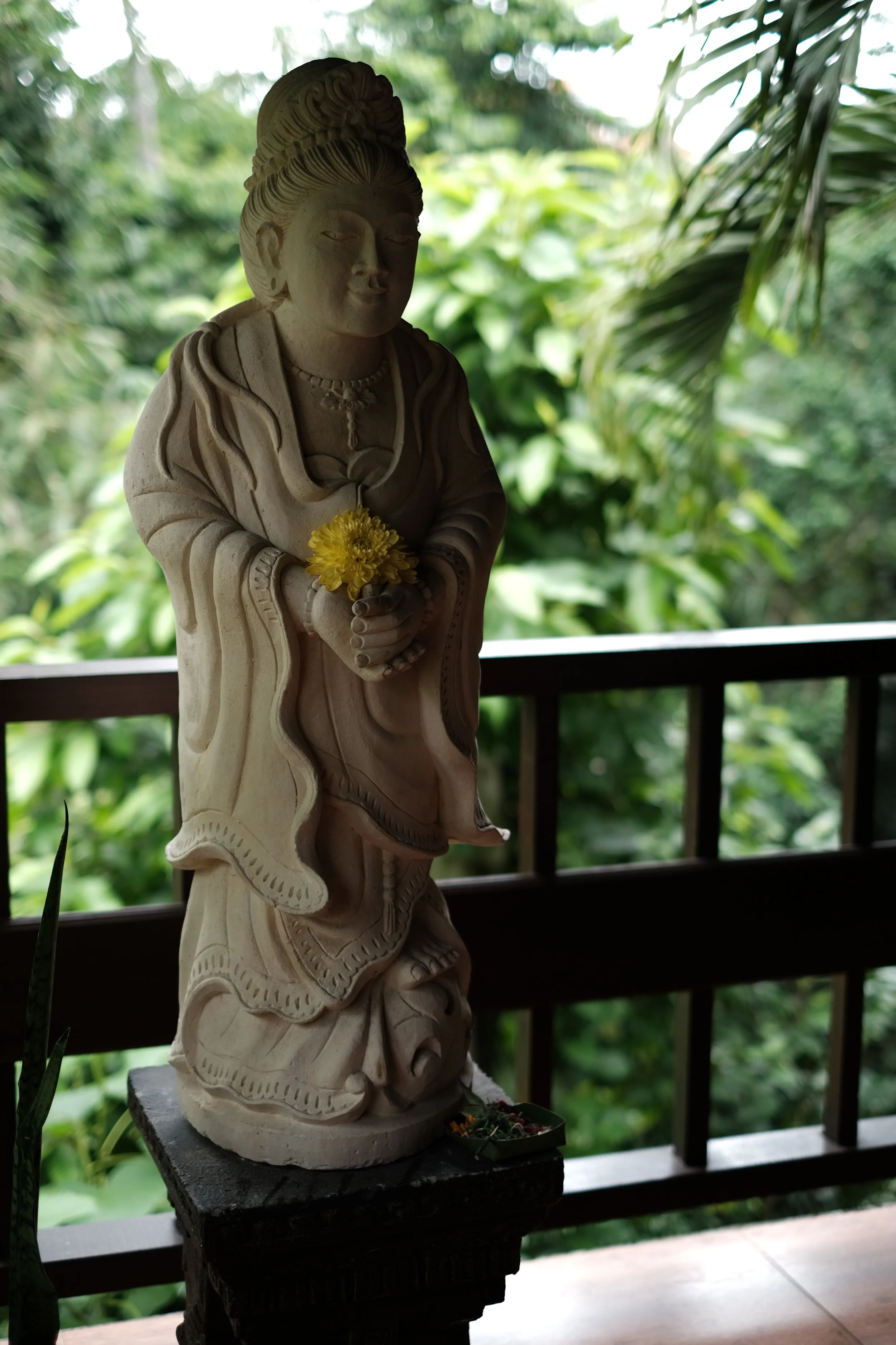 A carved stone statue on a balcony with lush green foliage in the background provides a wonderful reminder of the beauty of Bali.