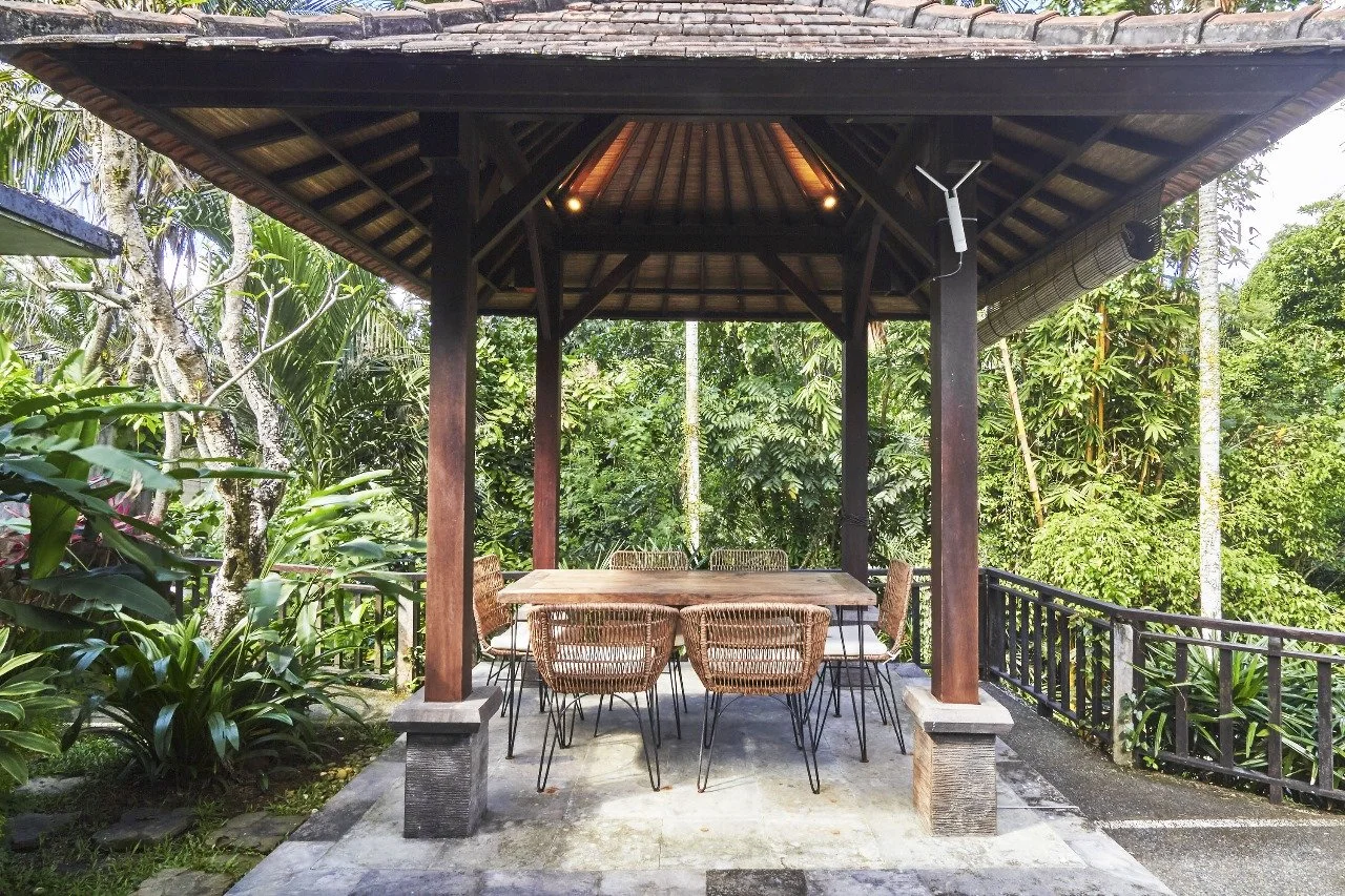 An outdoor sitting area with a wooden table and wicker chairs under a traditional wooden gazebo, surrounded by lush green tropical plants and trees.