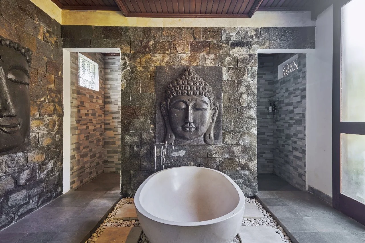 Interior of a bathroom with a freestanding bathtub in front of a textured Buddha face sculpture. To the left and right of the sculpture are toilet and shower areas with tiled walls.