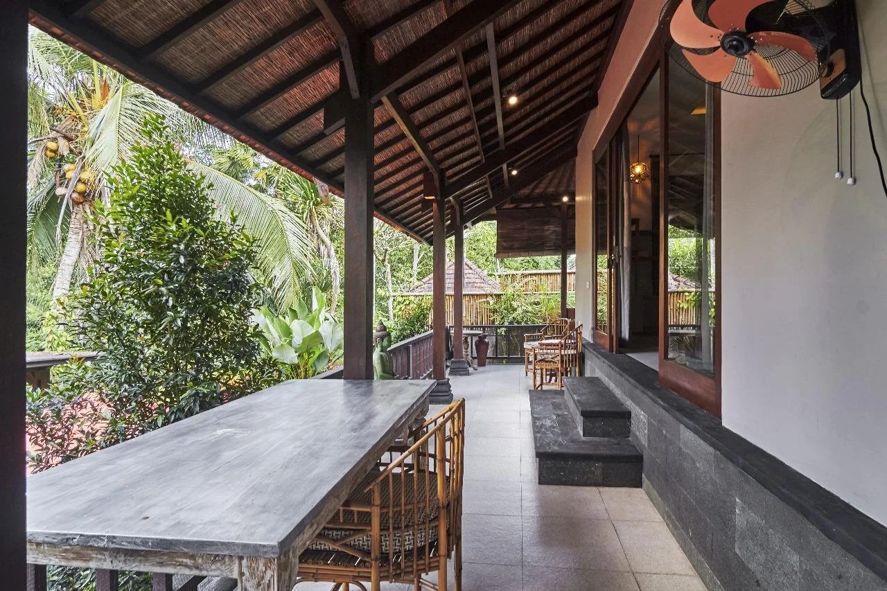 Balcony area with wooden furniture and lush tropical plants, overlooking a garden, with a ceiling fan, and sliding glass doors leading inside.