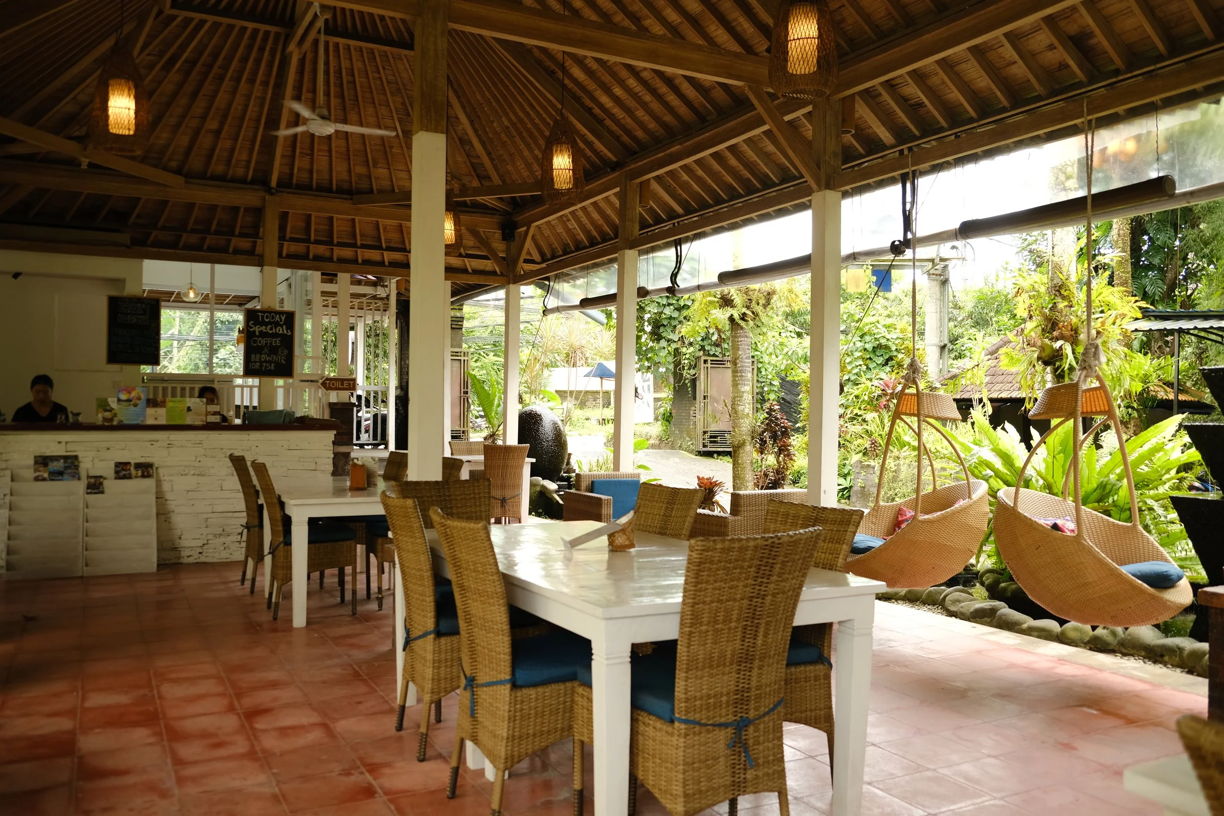 Open-air tropical cafe/restaurant with wicker chairs around white tables, hanging swing seats, lush greenery outside, and a wooden ceiling.