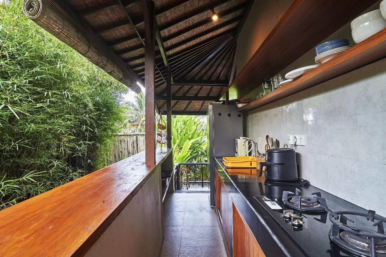 Outdoor kitchen with a black countertop, gas stove, toaster, wooden shelves holding dishes and glasses, surrounded by lush green plants and bamboo.