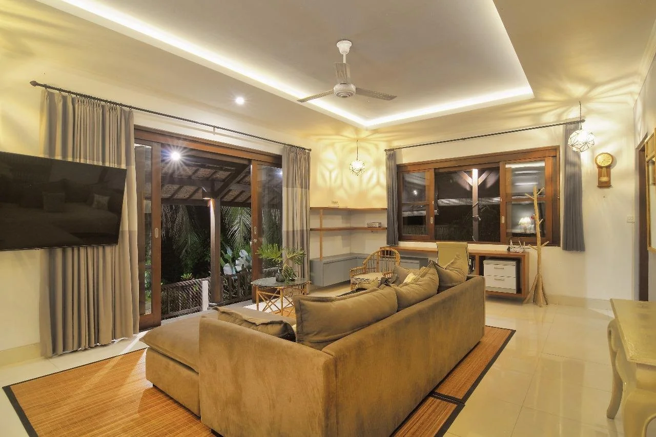 Living room with sofa, large windows with curtains, sliding glass door, mounted TV, ceiling fan, hanging light fixtures, and outdoor patio visible through sliding door.