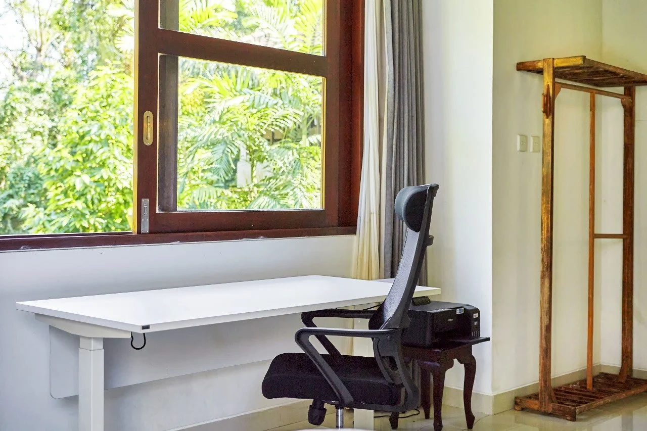 An office corner with a white desk, black ergonomic office chair, window with a view of lush green trees makes for a great place to study or work.