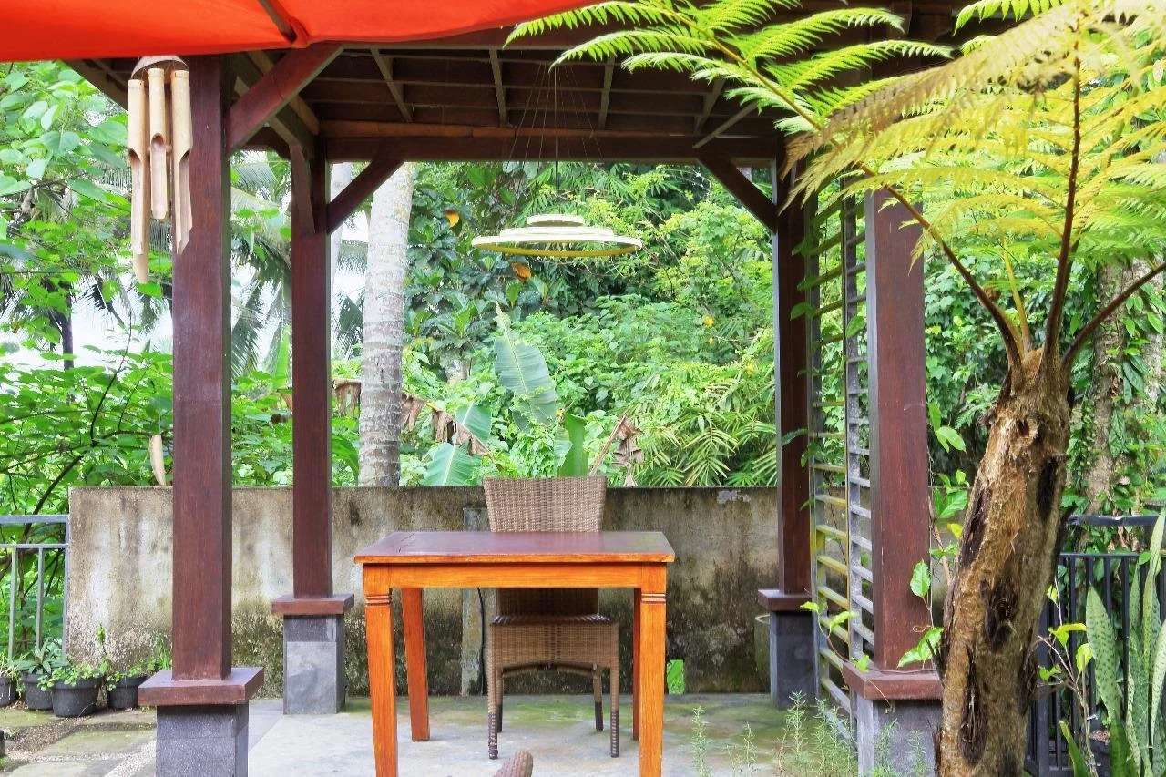 Outdoor shaded patio area with wooden table and wicker chair, surrounded by lush green tropical plants and trees.