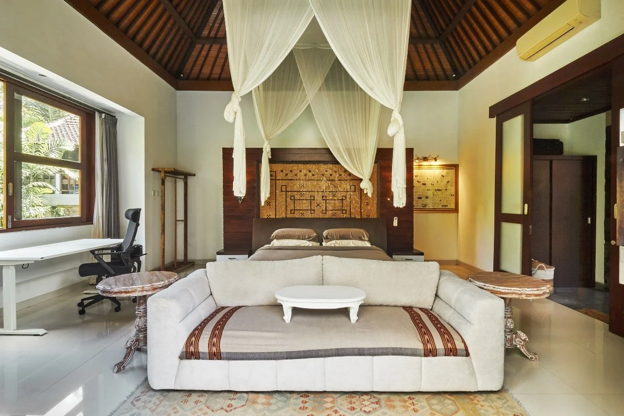 Luxury bedroom with a canopy bed, white sofa, wooden side tables, and large windows in a tropical-style room.