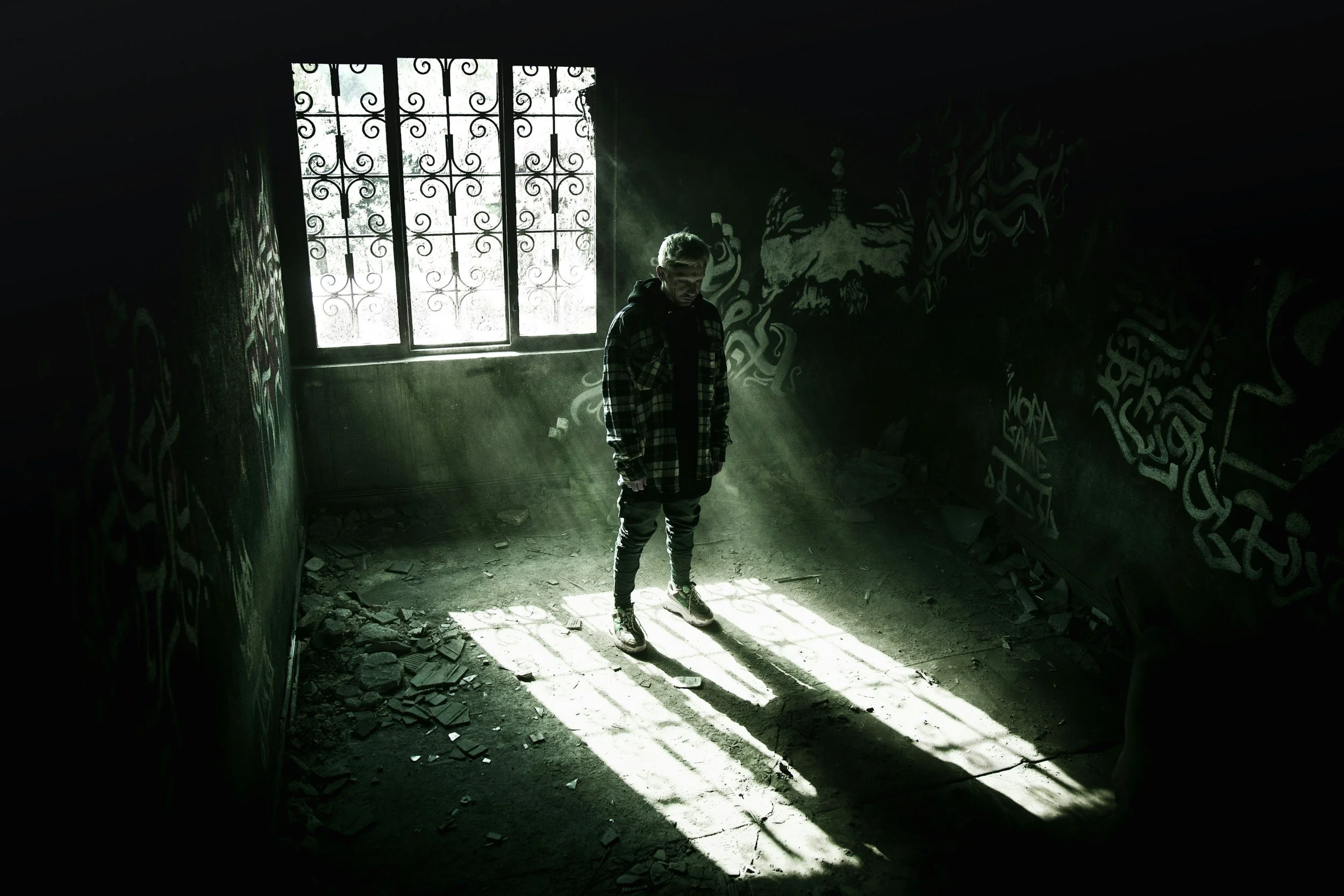 A young man stands alone in a dark, abandoned room with graffiti-covered walls, illuminated by sunlight streaming through a barred window, casting shadows on the floor.