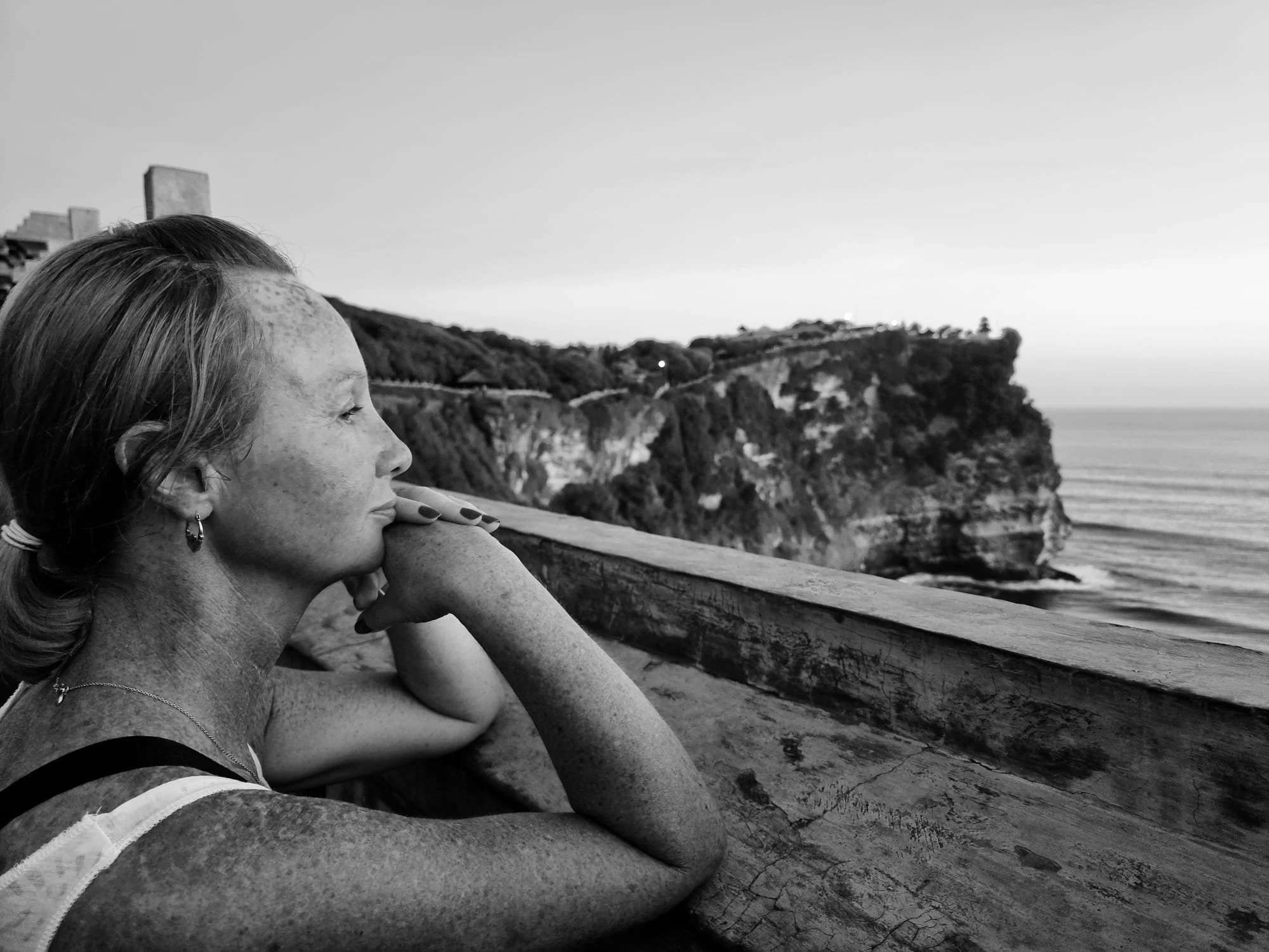 A woman with long hair and freckles rests her head on her hand, looking towards a landscape of cliffs and ocean on a clear day.