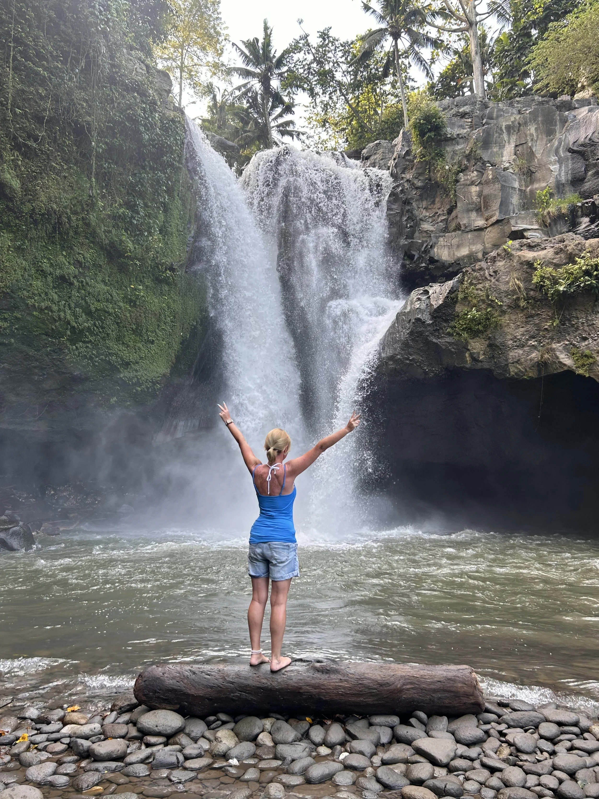 A woman standing on a log at the edge of a river, facing a waterfall with lush green trees and rocks in the background, arms raised in celebration.