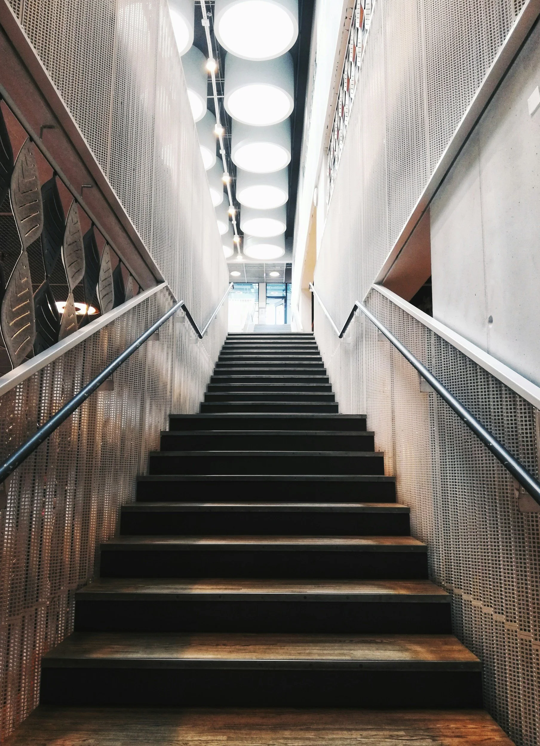 Indoor staircase with black steps, metal handrails, metal perforated walls, circular ceiling lights, and a glass door at the top.