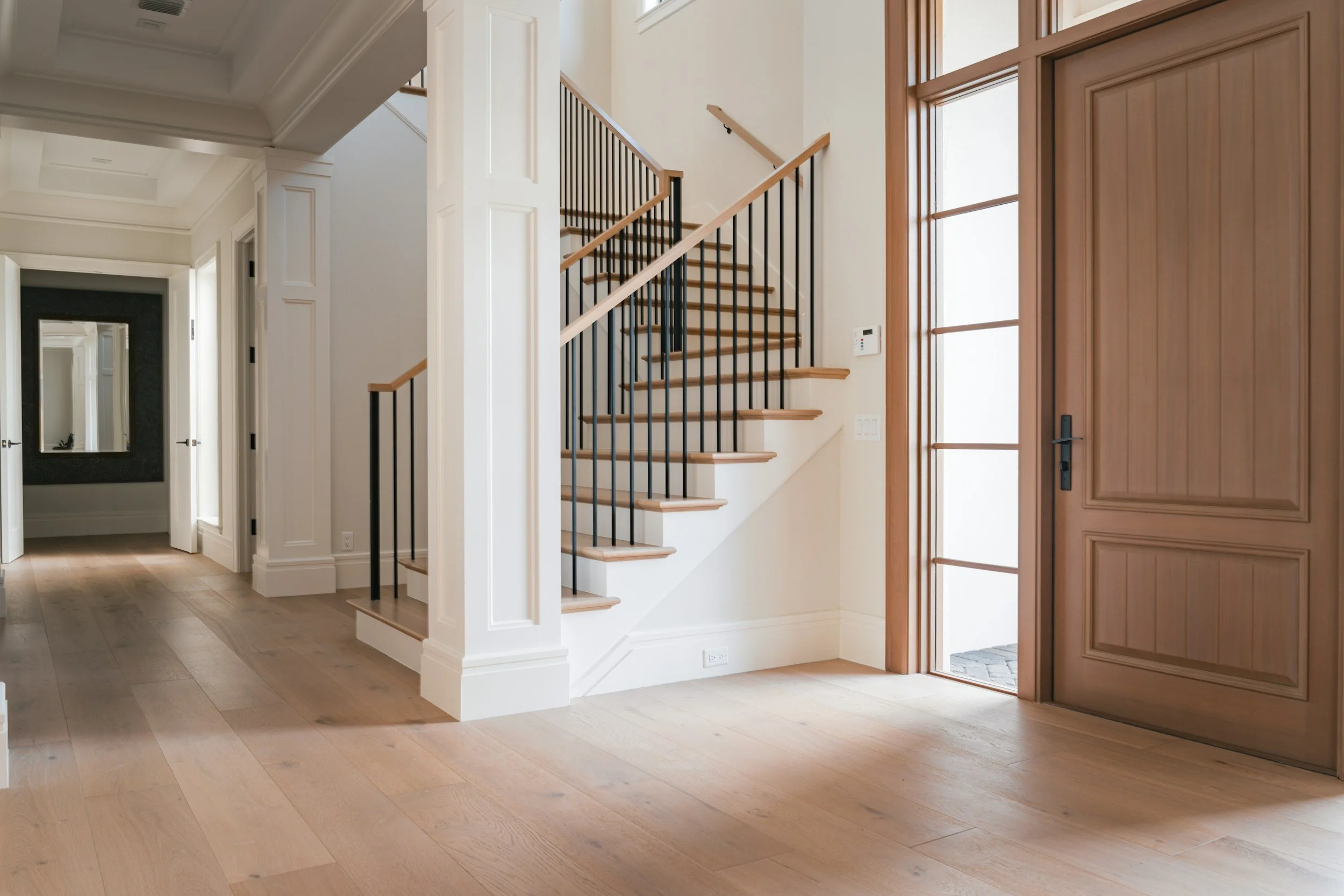 Interior of a modern home with a staircase featuring black railings and wooden steps, next to a large wooden door, in a well-lit entryway with light-colored hardwood floors and white walls.