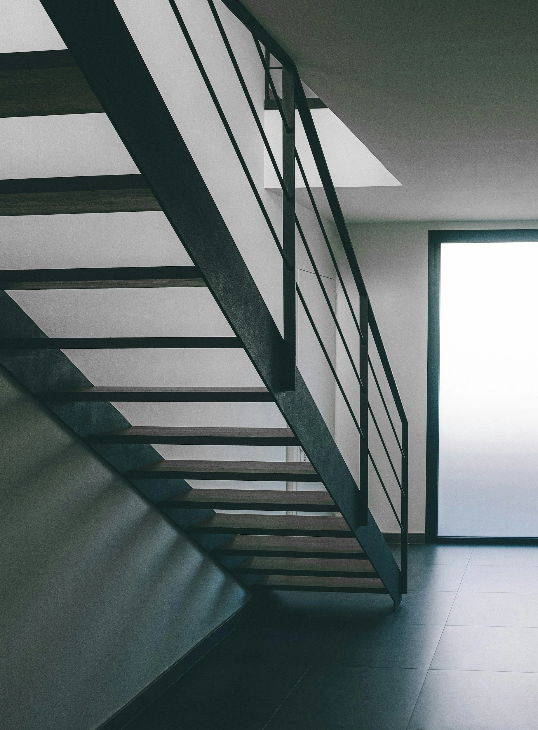 Interior view of a modern stairway with wooden steps and black metal railing, next to a window allowing natural light to illuminate the space.