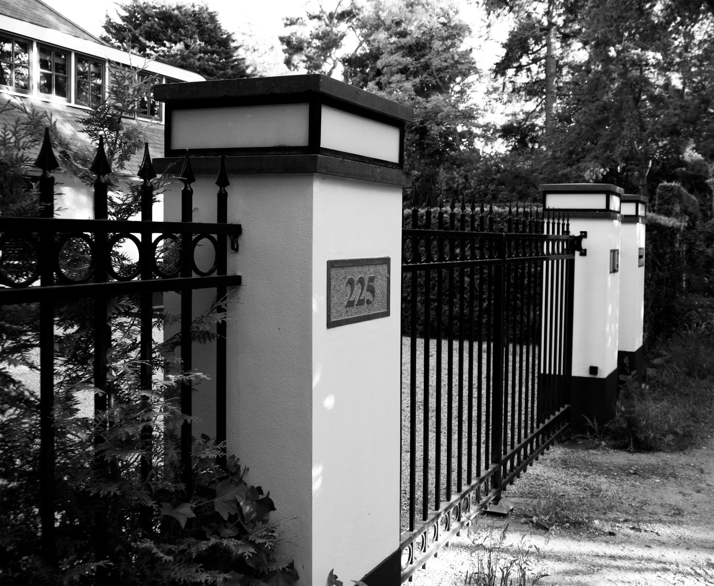 Black and white photo of a modern fence with white posts and black metal railing, surrounded by trees and plants, with a house visible in the background.