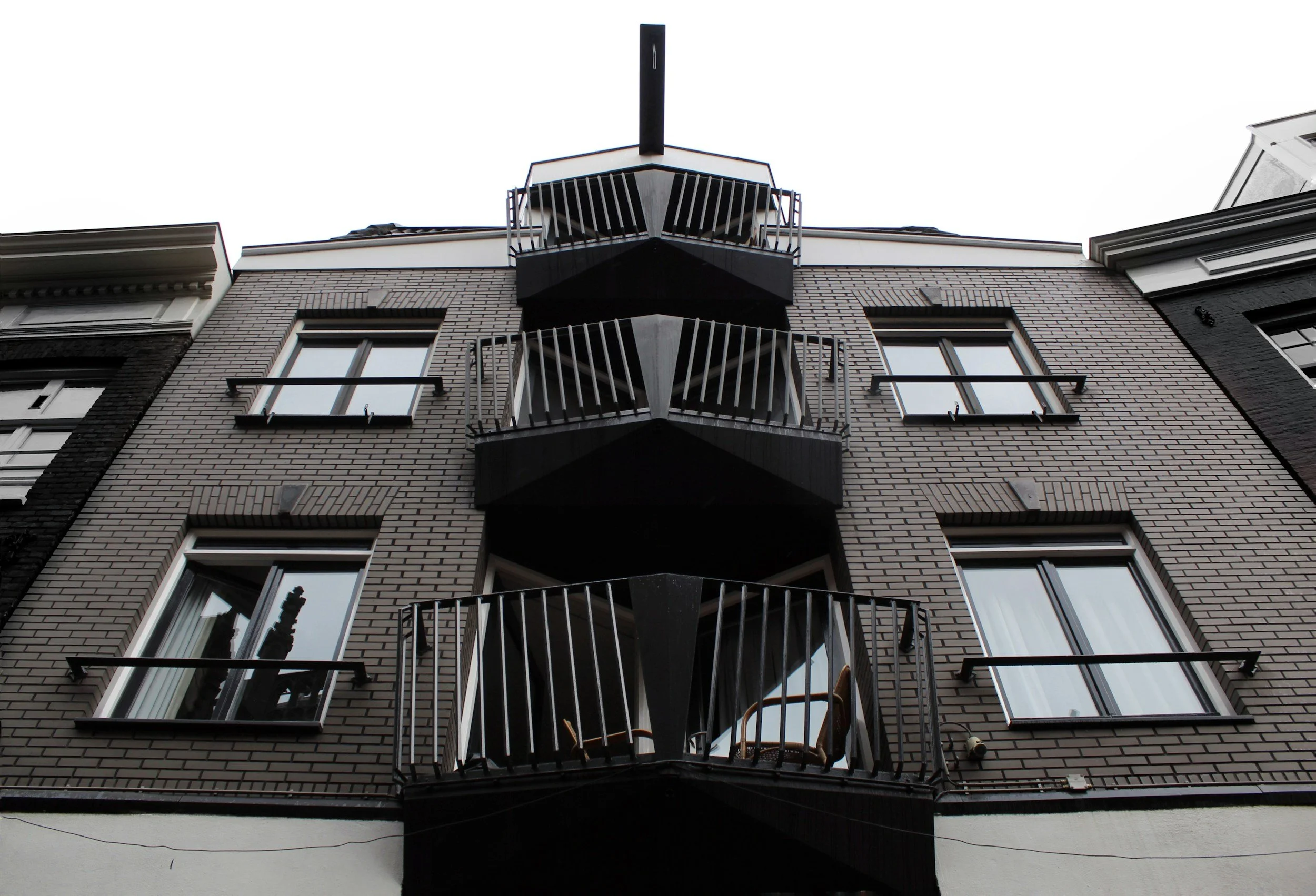 A view of a multistory brick building with six balconies, four on the bottom and two on the top, with black metal railings, and windows with white frames, against a bright overcast sky.