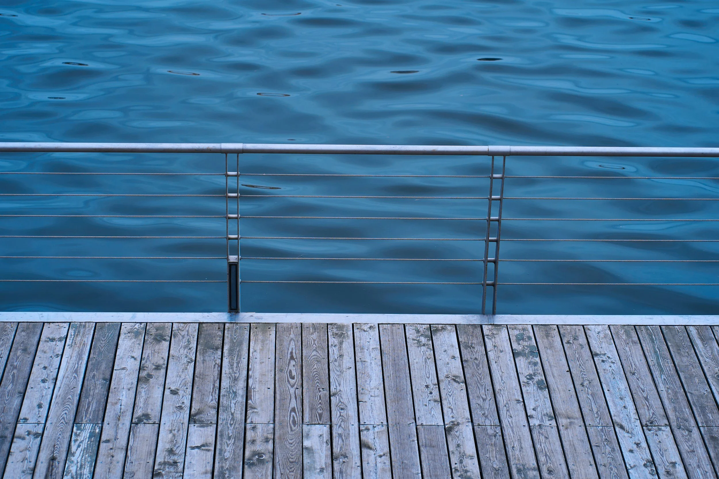 View of calm blue water from a dock with wooden planks and a metal railing.