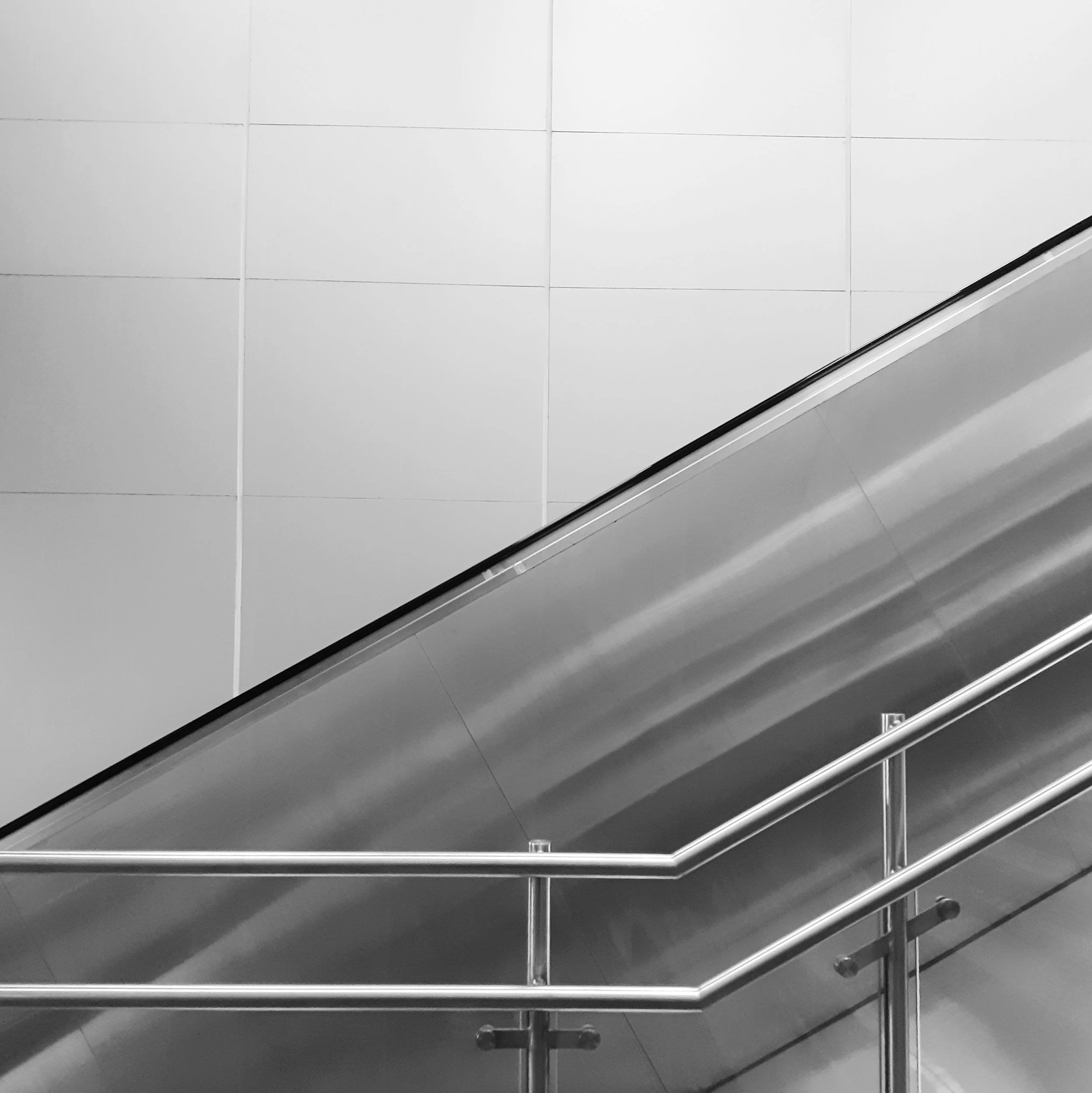The image shows a section of a modern staircase with metallic handrails and large white wall tiles in the background.