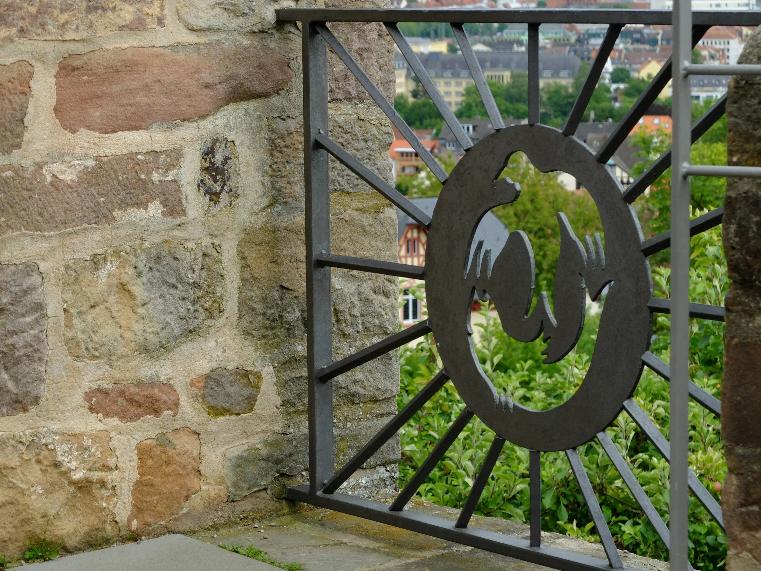 Close-up view of a stone wall with a decorative metal sculpture of a horse's head integrated into a metal gate, overlooking a cityscape with various buildings and greenery in the background.