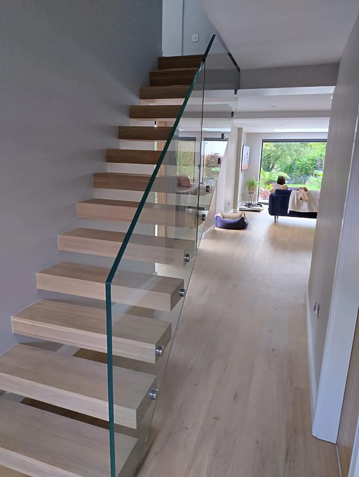 Modern home interior with wooden floating staircase and glass railing leading to living room with large window, woman sitting on sofa, and greenery outside.