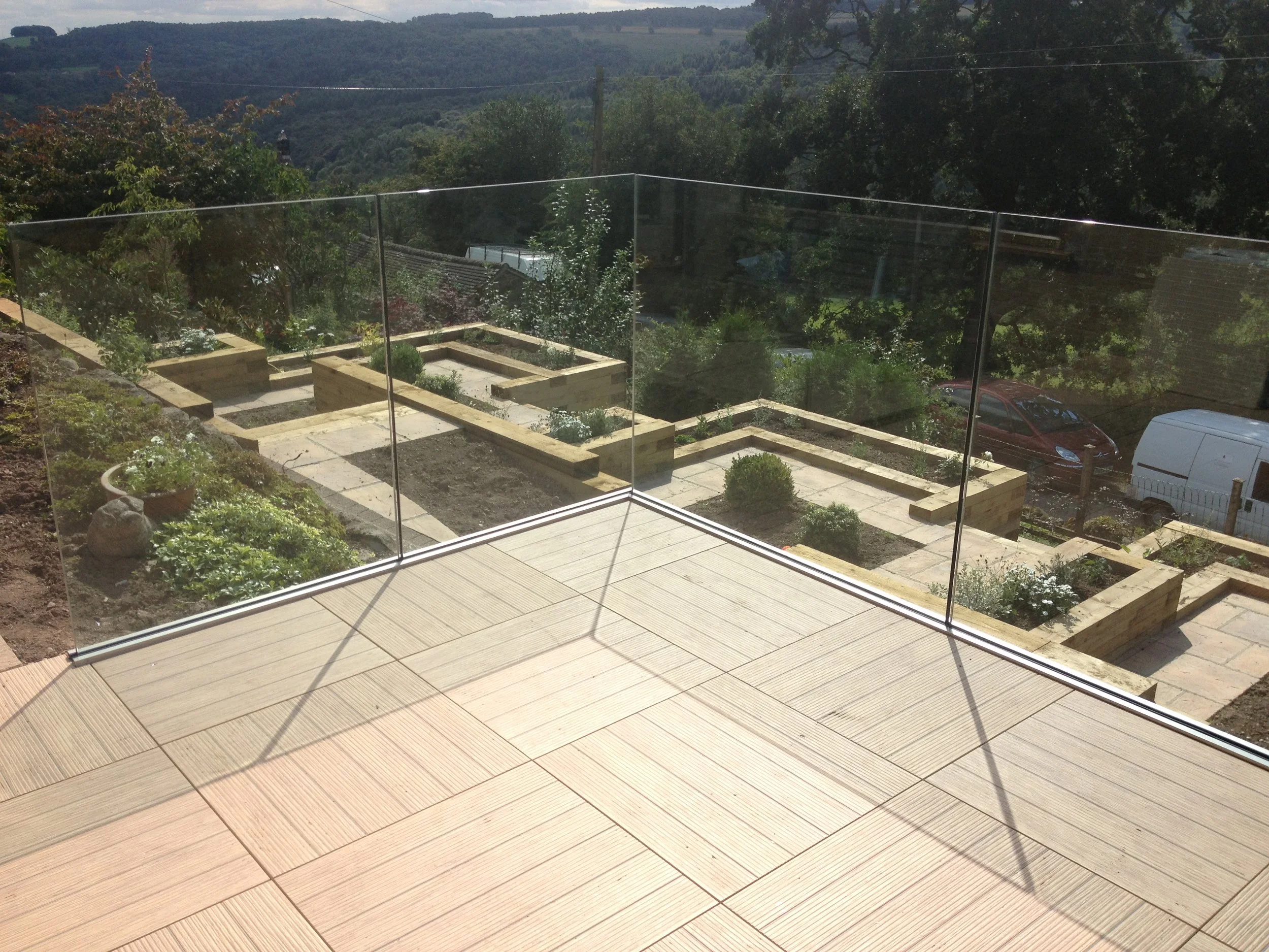 A view from a patio or balcony with light-colored wooden flooring, enclosed by glass panels. In the background, there are landscaped garden beds with plants and bushes, a paved pathway, and a wire fence. Beyond the garden, there are trees and a scenic hilly landscape in the distance under a partly cloudy sky.