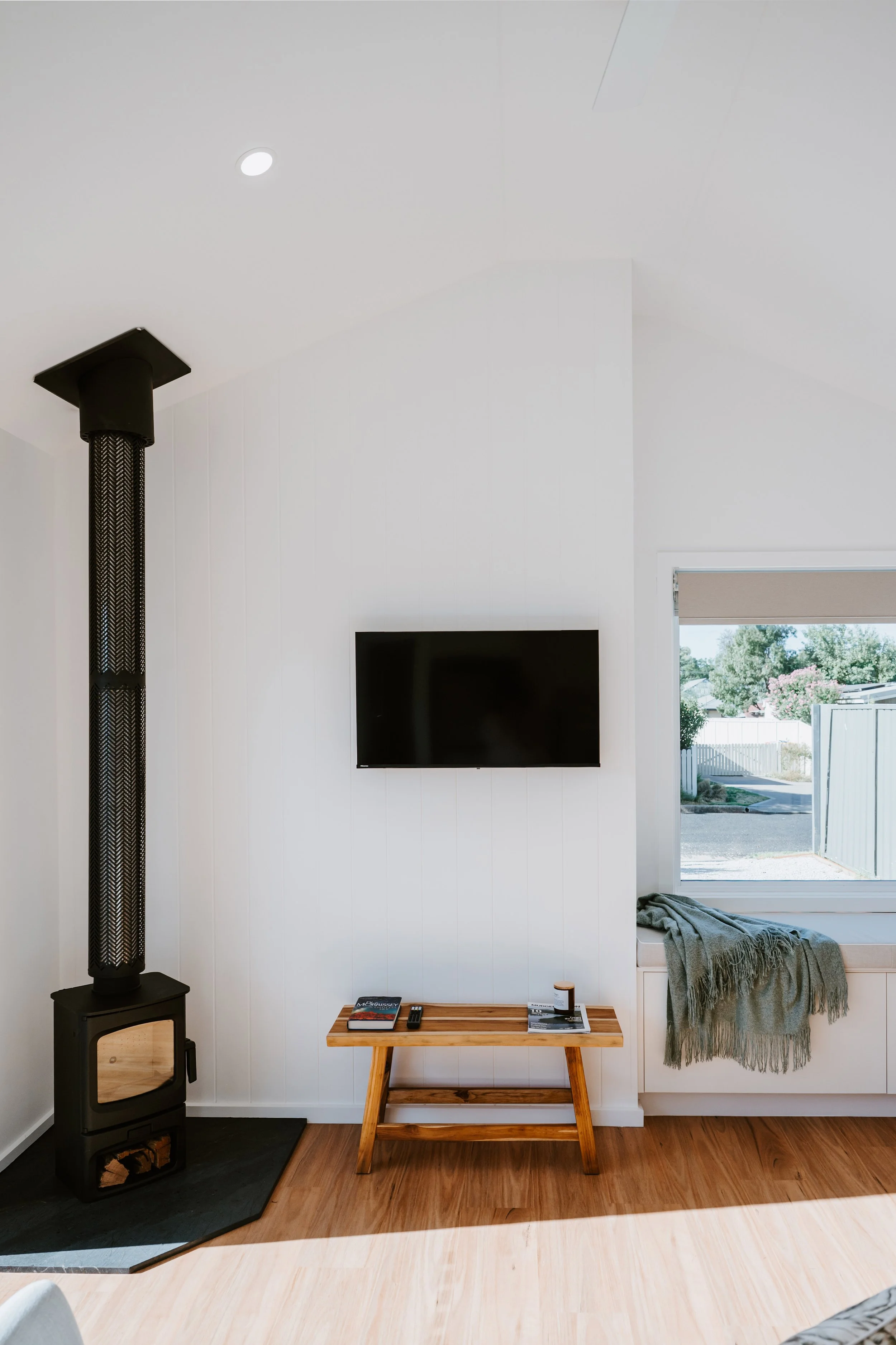 Living room with a wood stove on a black hearth, a TV mounted on a white wall, a small wooden table with magazines and remote, and a window with a view outside.