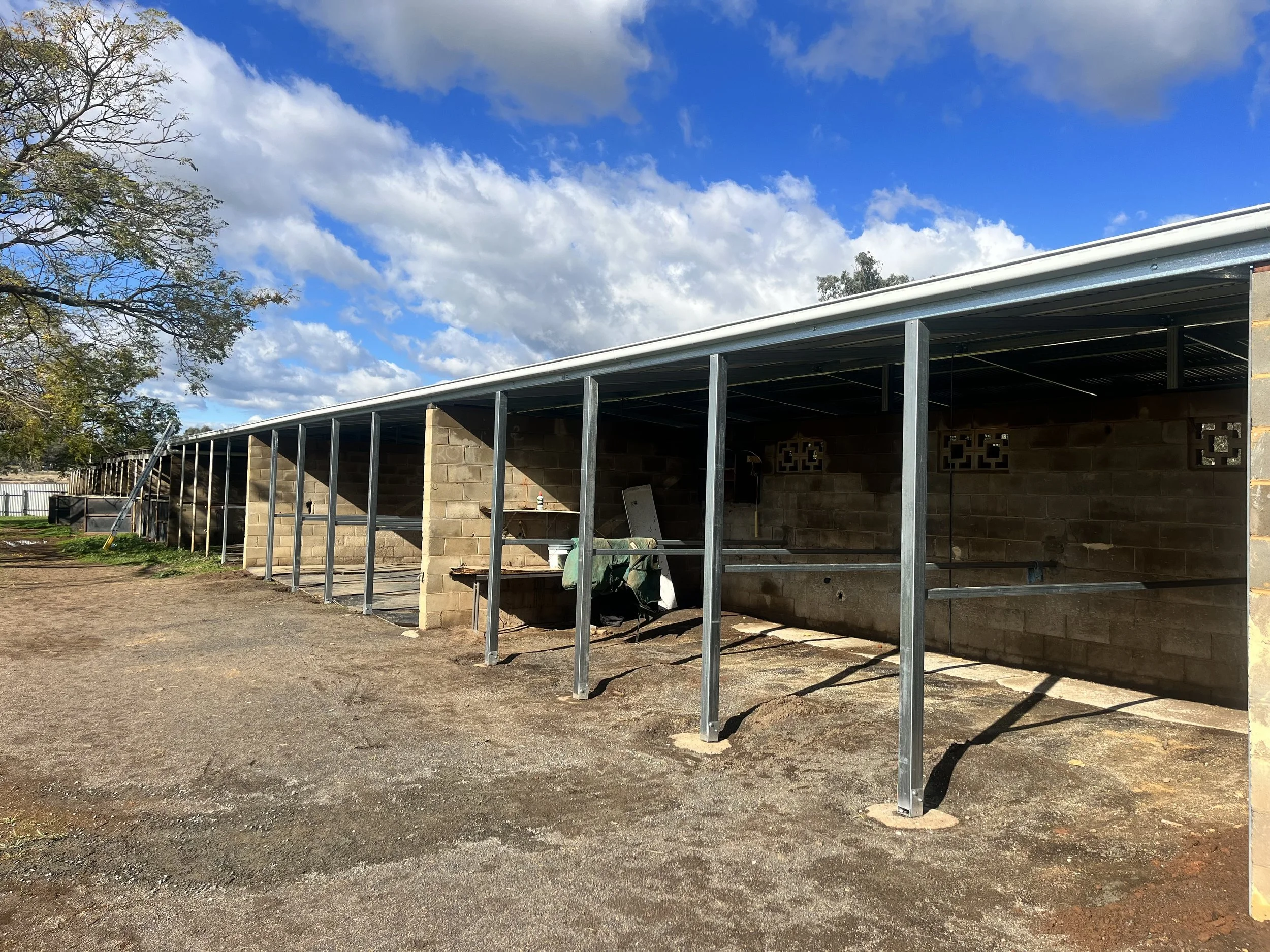 Construction site with a building framework of metal beams and brick walls, partly covered by a roof, under a partly cloudy blue sky, with a tree on the left side.