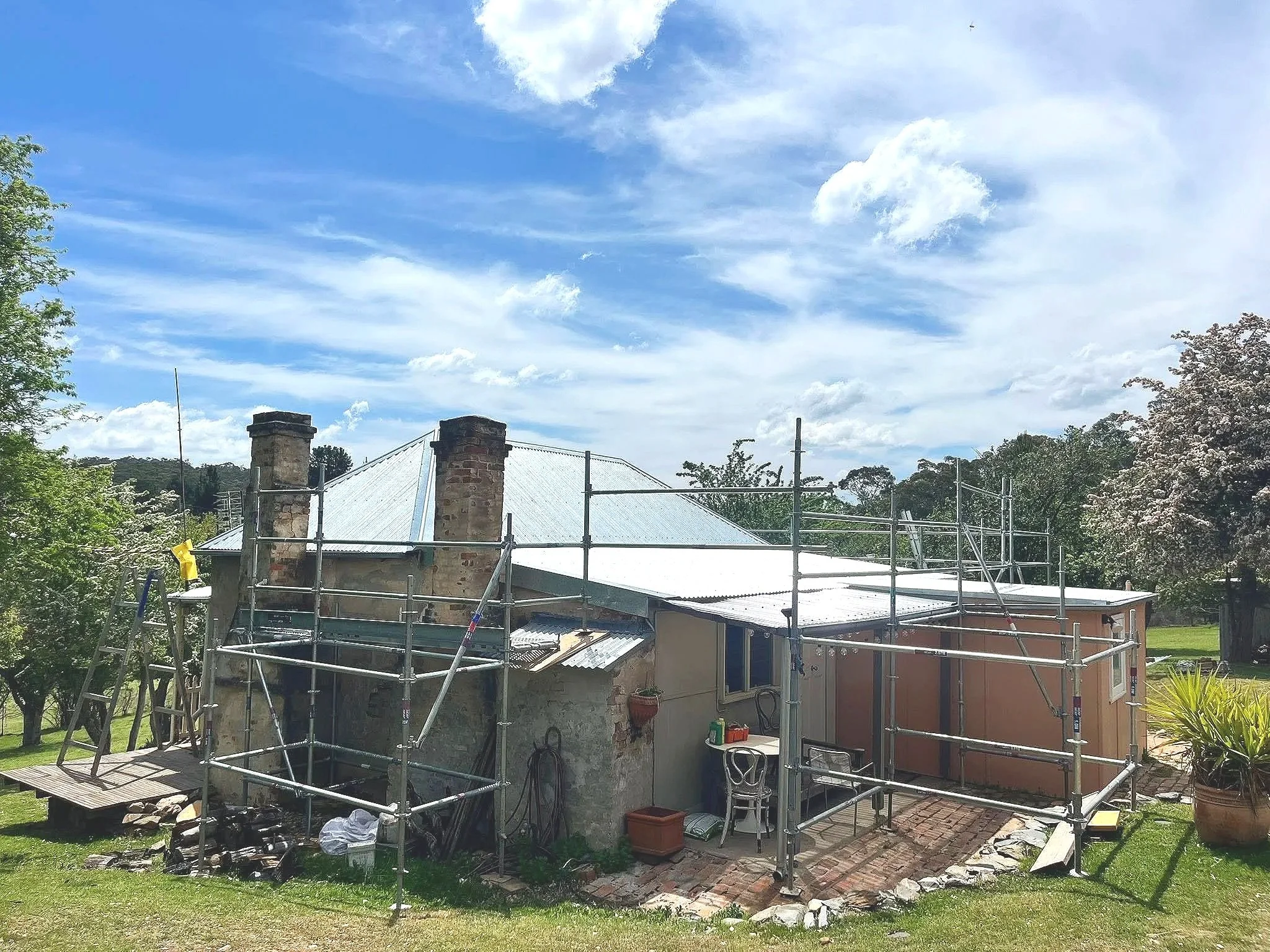A house under renovation or construction with scaffolding around it. The house has a brick chimney, metal roofing, and is surrounded by greenery and trees with a blue sky and clouds overhead.
