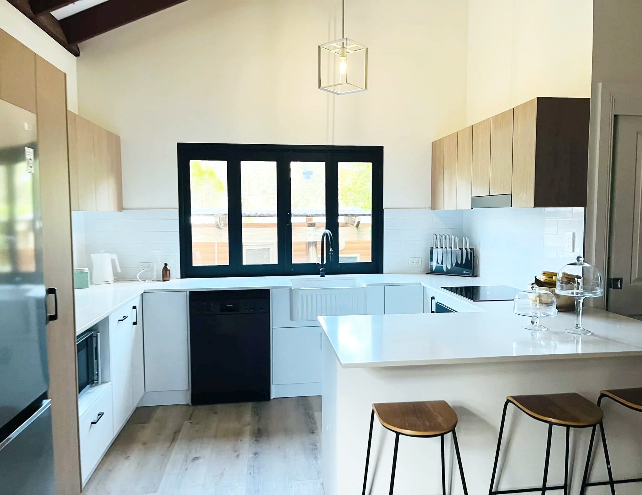 A modern kitchen with white cabinets, black window frames, and wooden chairs around a white island. The kitchen has a black stove, a kettle, and a knife holder on the countertop. The ceiling is sloped with wooden beams, and there is a hanging geometric light fixture. Large windows let in natural light and show greenery outside.