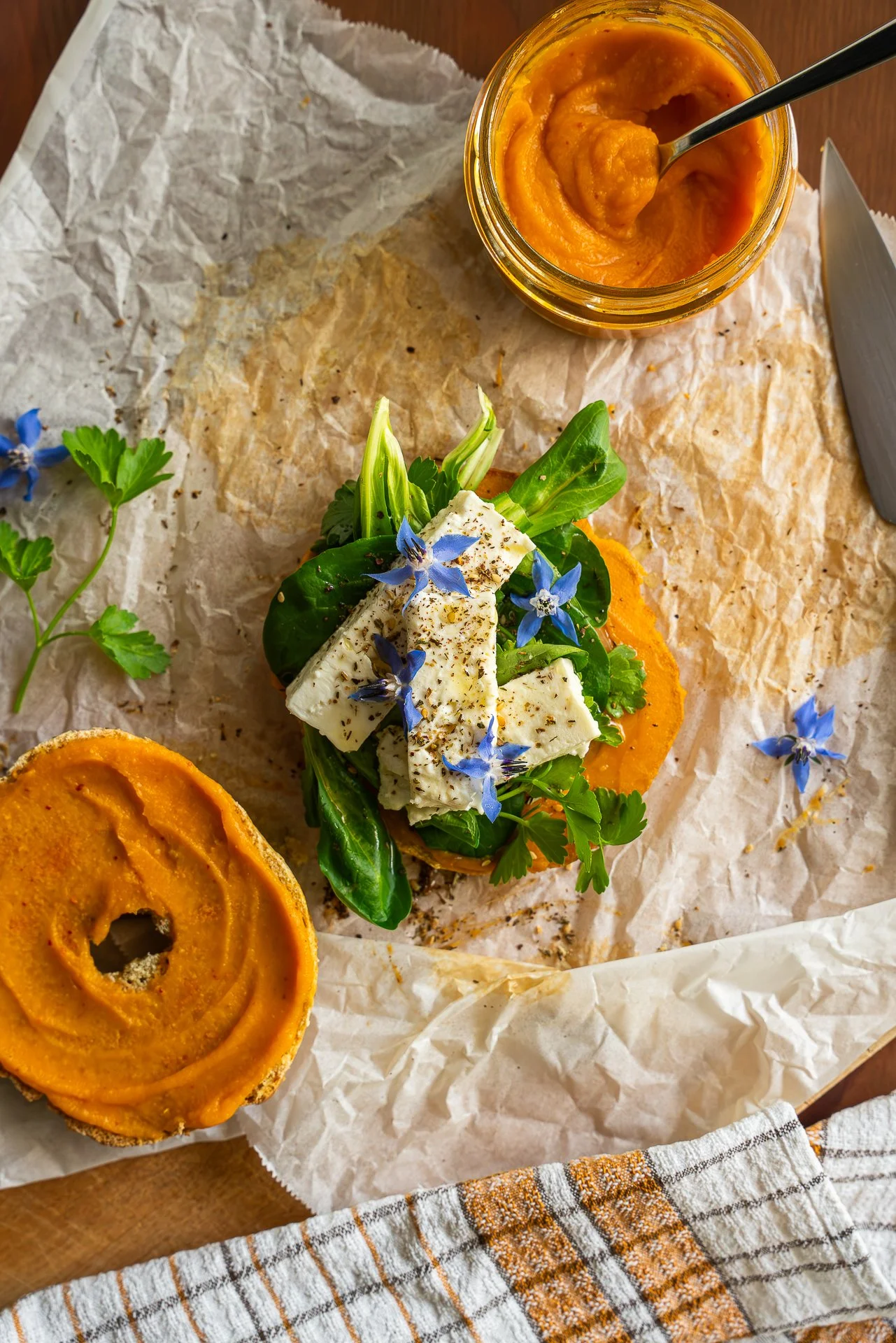 Bagel au zaatar, ouvert, garni d'herbes fraîches, de feta, fleurs de bourrache, pickles d'oignons lactofermentés, huile d'olive, tartinable à base de patate douce, yuzu et piment d'Espelette, le tout posé sur du papier sulfurisé. Vue du dessus.