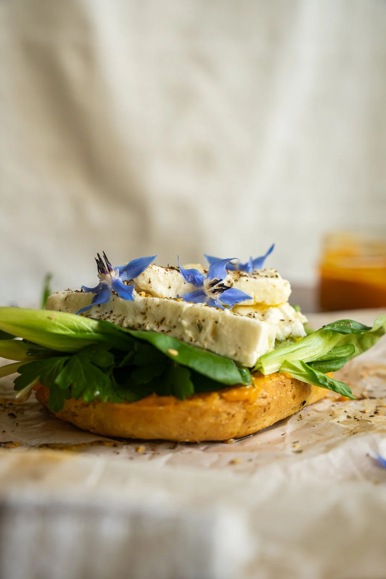 Bagel au zaatar, ouvert, garni d'herbes fraîches, de feta, fleurs de bourrache, pickles d'oignons lactofermentés, huile d'olive, tartinable à base de patate douce, yuzu et piment d'Espelette, le tout posé sur du papier sulfurisé.