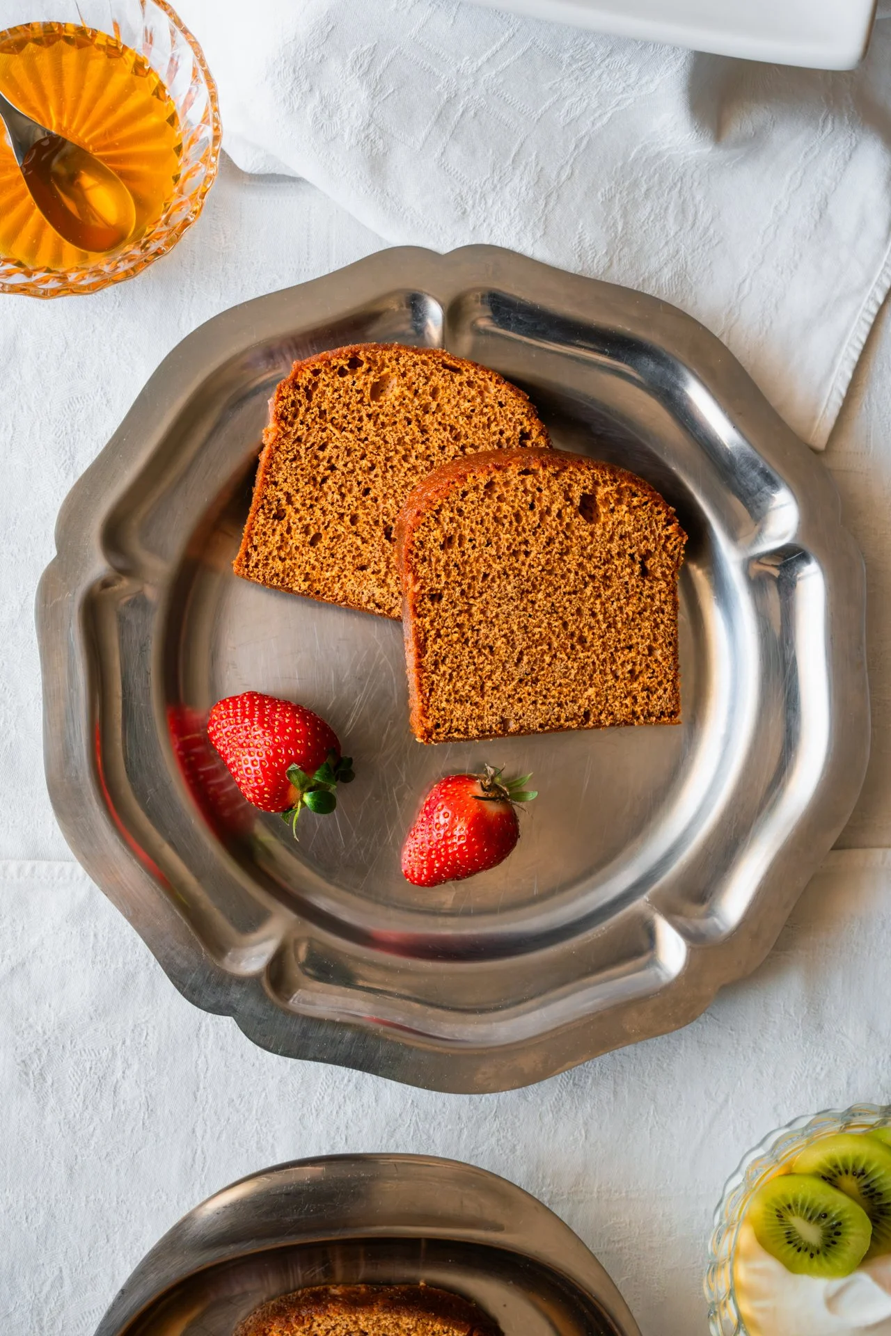 Deux tranches de pain d'épices, deux fraises fraiches sur un plateau en métal, reposant sur une table recouverte d'une nappe blanche, une coupelle en verre remplie de miel et une autre remplie de fromage blanc et de tranches de kiwi.