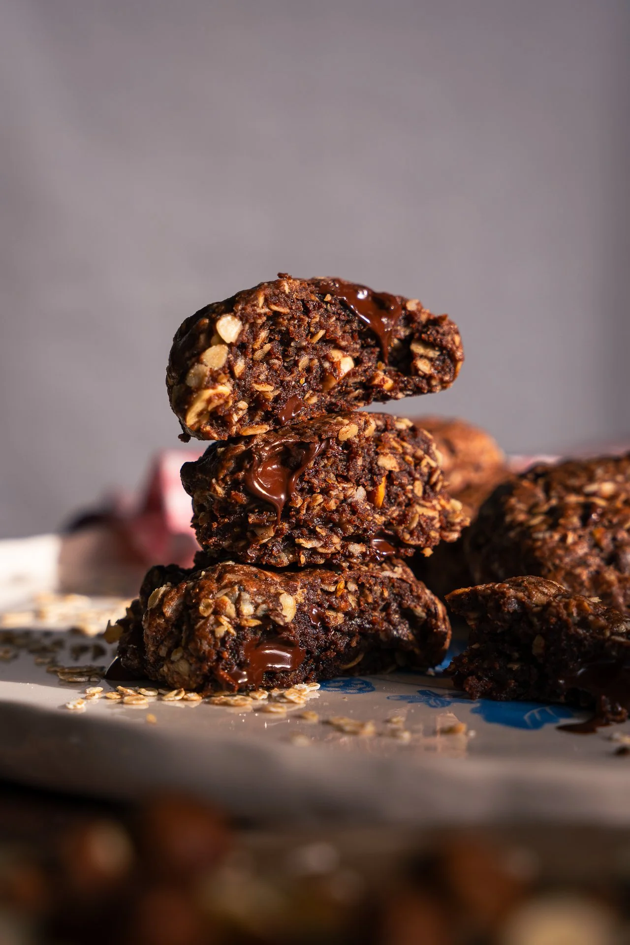 Cookies aux noisettes, avoine et chocolat noir coulant, empilés sur une assiette, avec des flocons d'avoine et des noisettes autour.