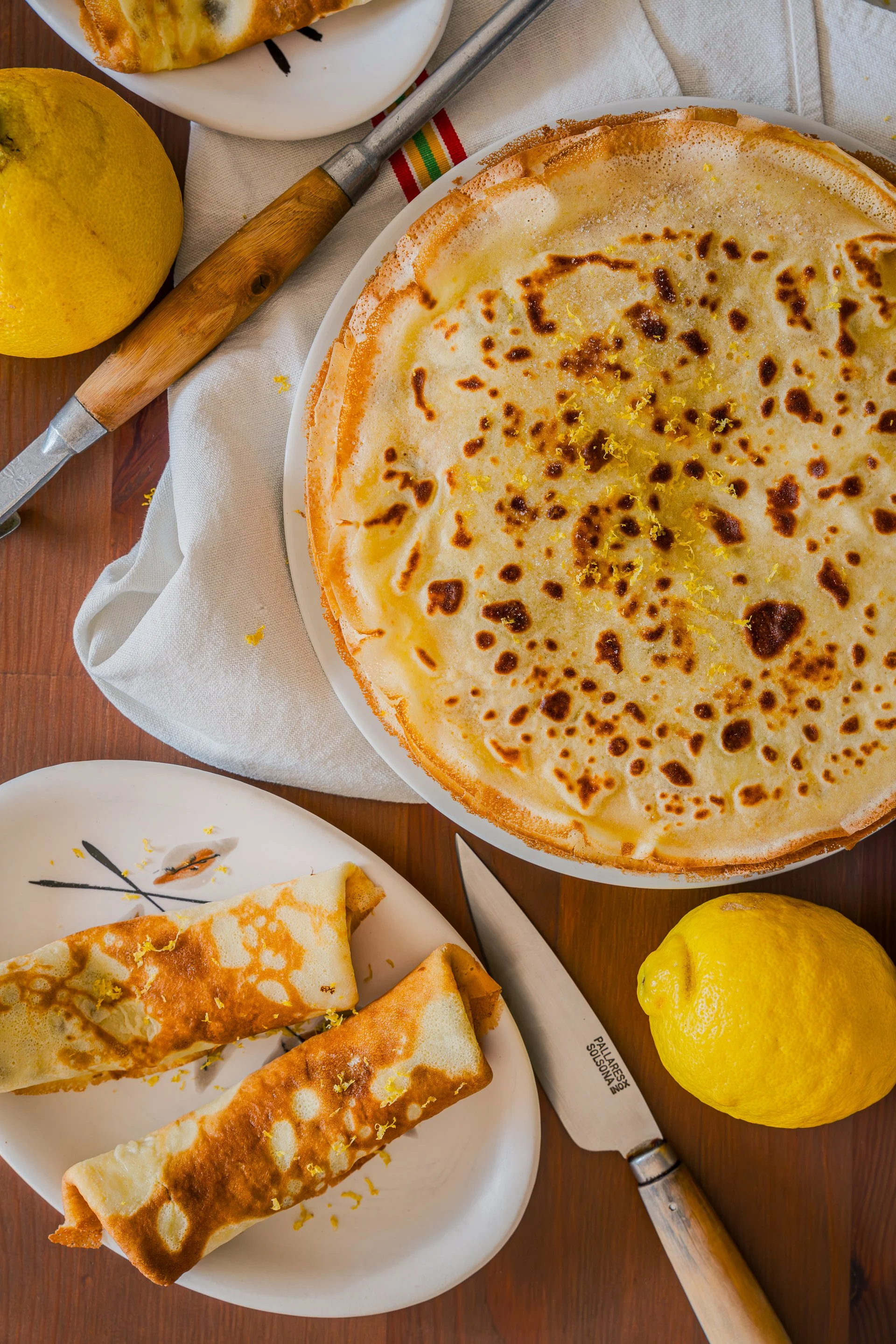 Crêpe garnie de zestes de citron bergamote et de sucre, deux crêpes roulées, deux citrons, un couteau de cuisine, une louche, et une assiette avec d'autres crêpes, sur une table en bois, vue du dessus.