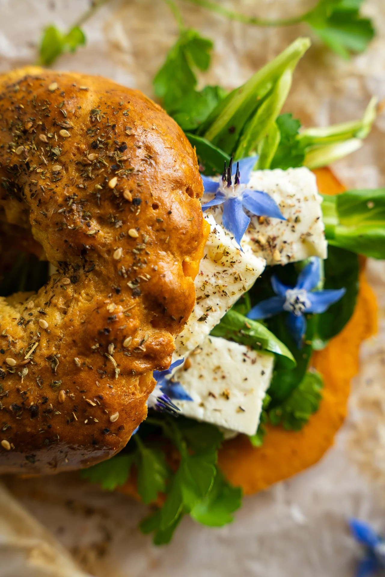 Bagel au zaatar, garni d'herbes fraîches, de feta, fleurs de bourrache, pickles d'oignons lactofermentés, huile d'olive, tartinable à base de patate douce, yuzu et piment d'Espelette. Vue du dessus, rapprochée.