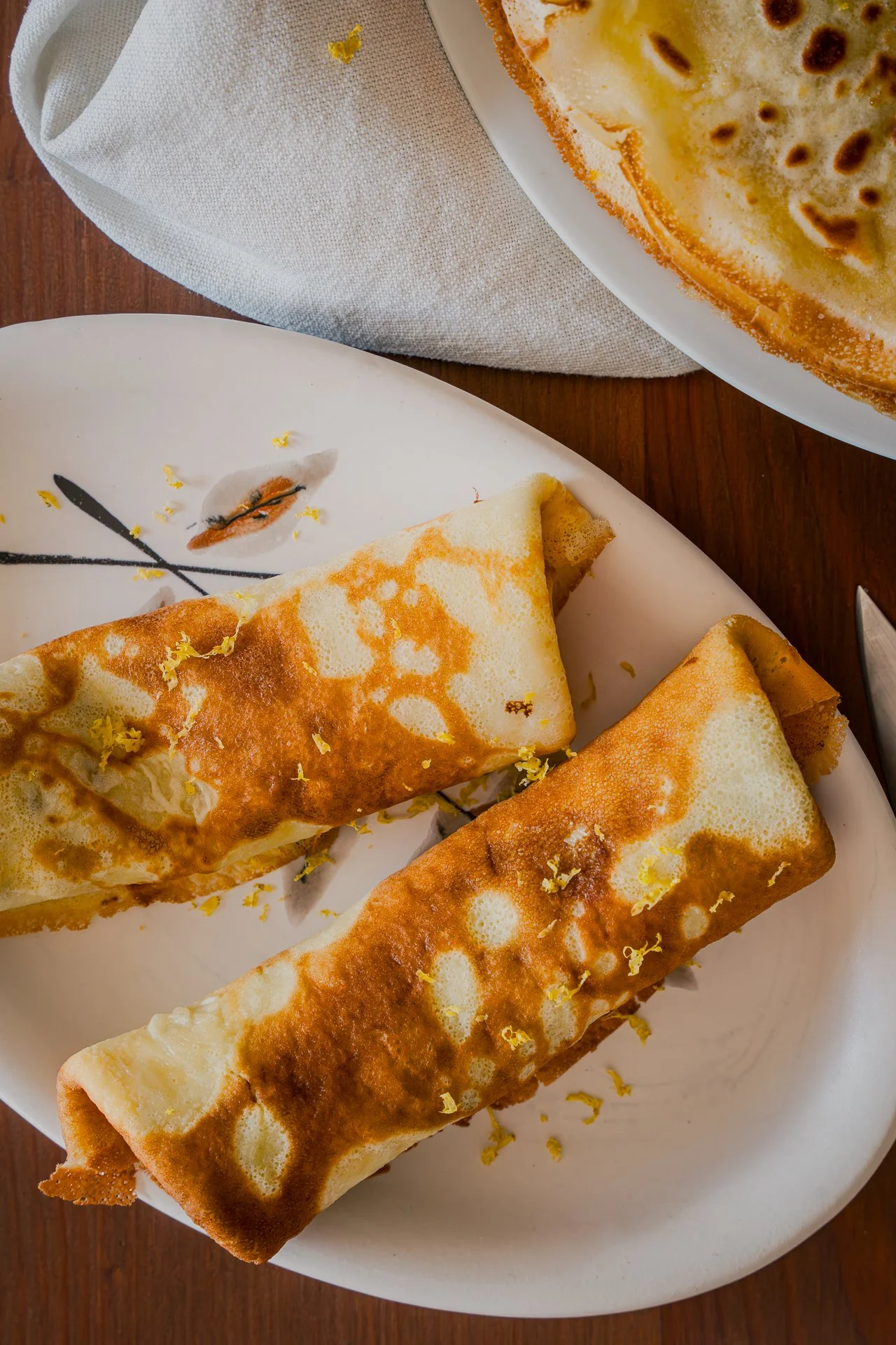 Deux crêpes roulées dans une petite assiette ovale et recouvertes de zestes de citron bergamote râpés, une autre assiette avec d'autres crêpes reposant sur un torchon, le tout sur une table en bois, vue du dessus.