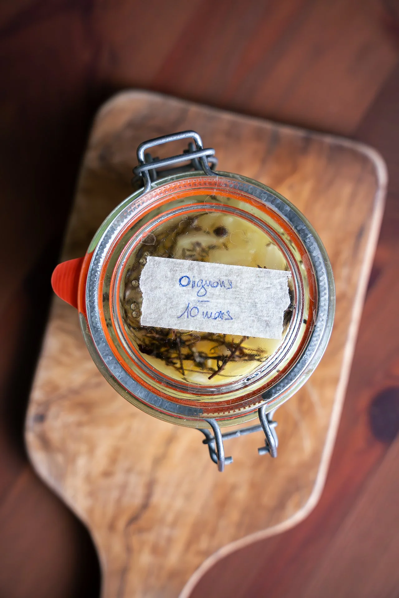 Pickles d'oignon jaune en bocal, avec branches de thym et romarin, vue du dessus, avec une étiquette indiquant 'Oignons 10 mars', posé sur une planche en bois.