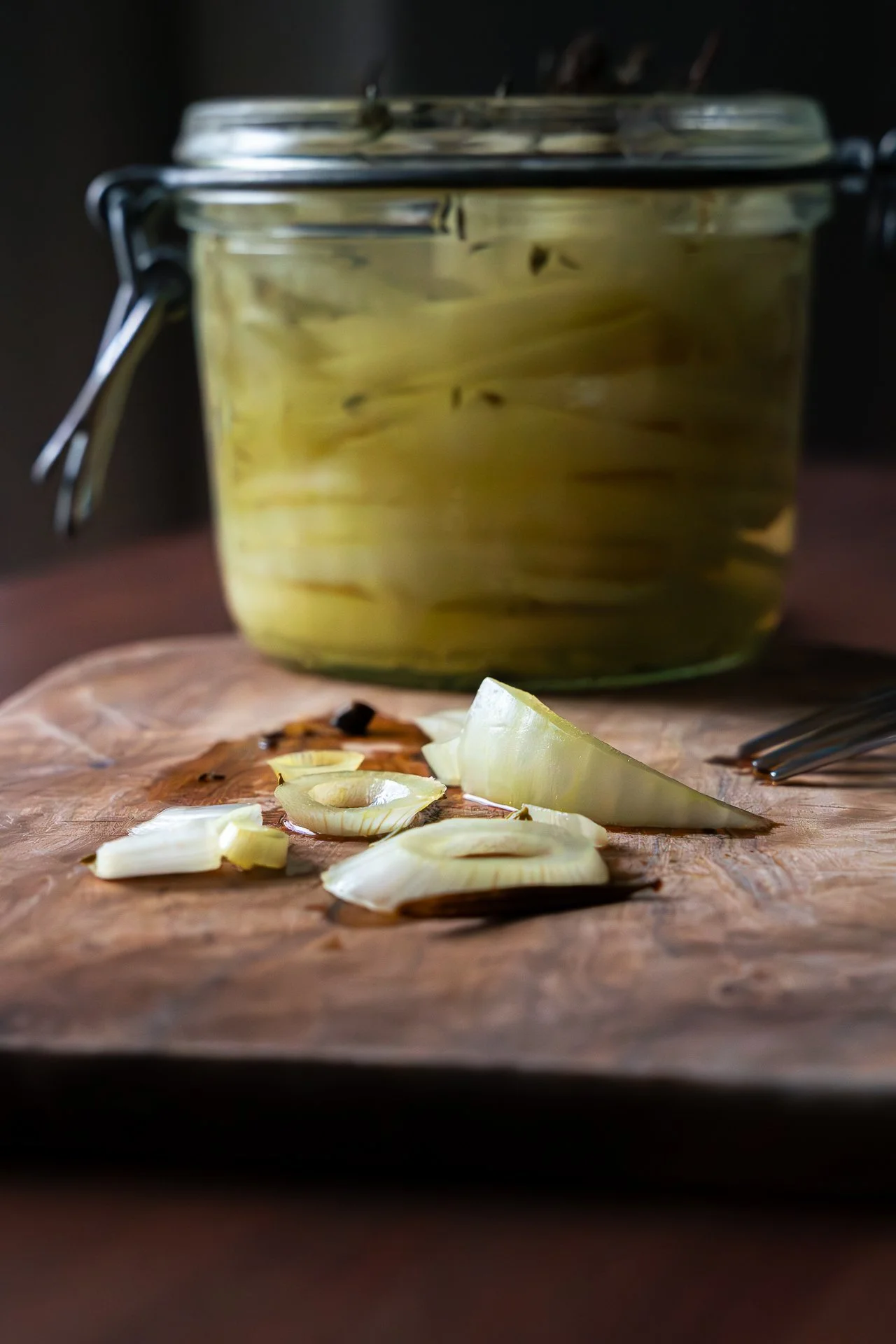 Pickles d'oignon jaune et une petite fourchette sur une planche en bois, devant un bocal en verre rempli de pickles en arrière-plan.
