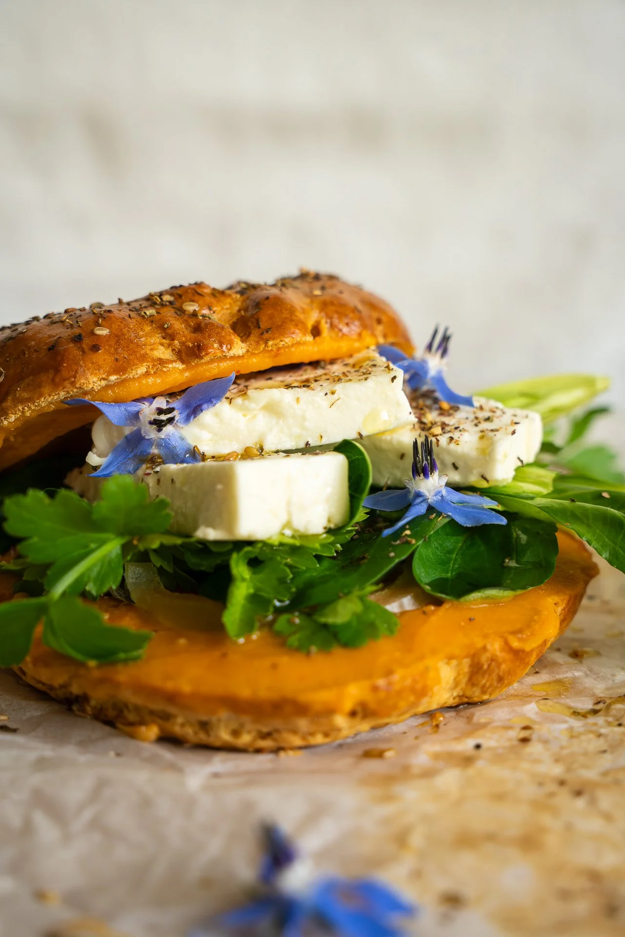 Bagel au zaatar garni d'herbes fraîches, de feta, fleurs de bourrache, pickles d'oignons lactofermentés, huile d'olive, tartinable à base de patate douce, yuzu et piment d'Espelette, le tout posé sur du papier sulfurisé.