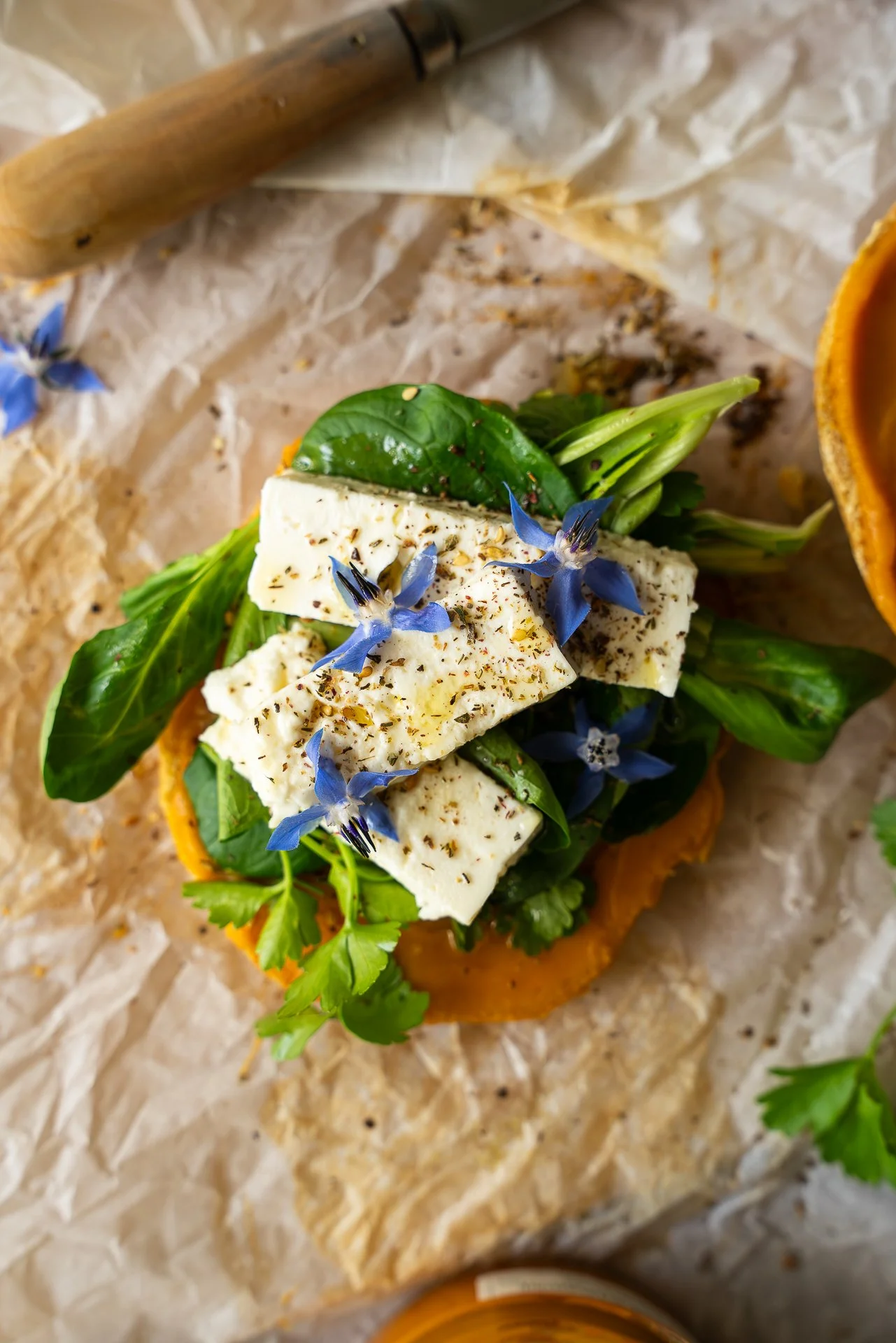 Bagel au zaatar, ouvert, garni d'herbes fraîches, de feta, fleurs de bourrache, pickles d'oignons lactofermentés, huile d'olive, tartinable à base de patate douce, yuzu et piment d'Espelette, le tout posé sur du papier sulfurisé. Vue du dessus.